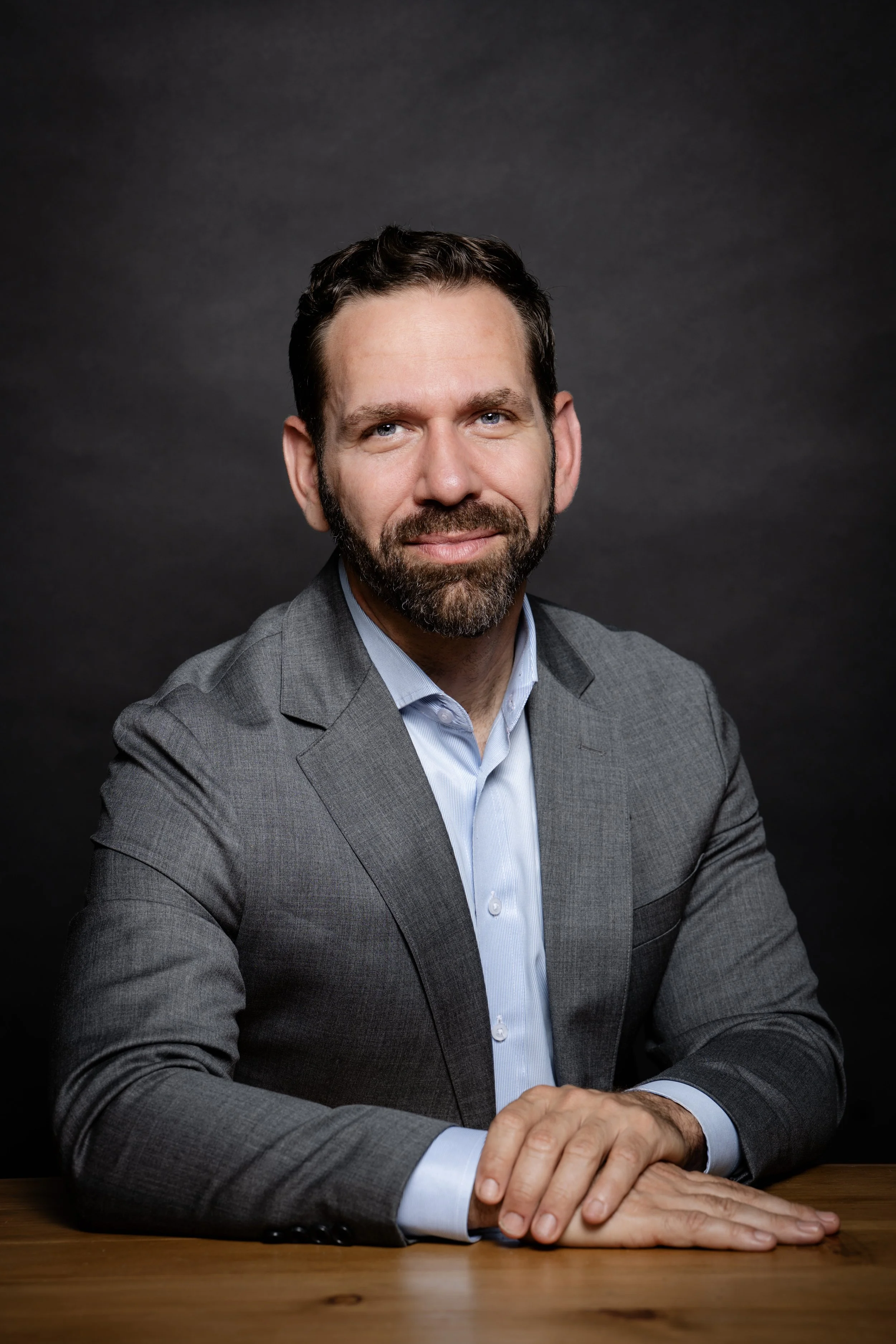 A man with a beard and short dark hair sitting at a wooden table, wearing a gray suit and light blue dress shirt, against a dark gray background for a corporate headshot done in a studio