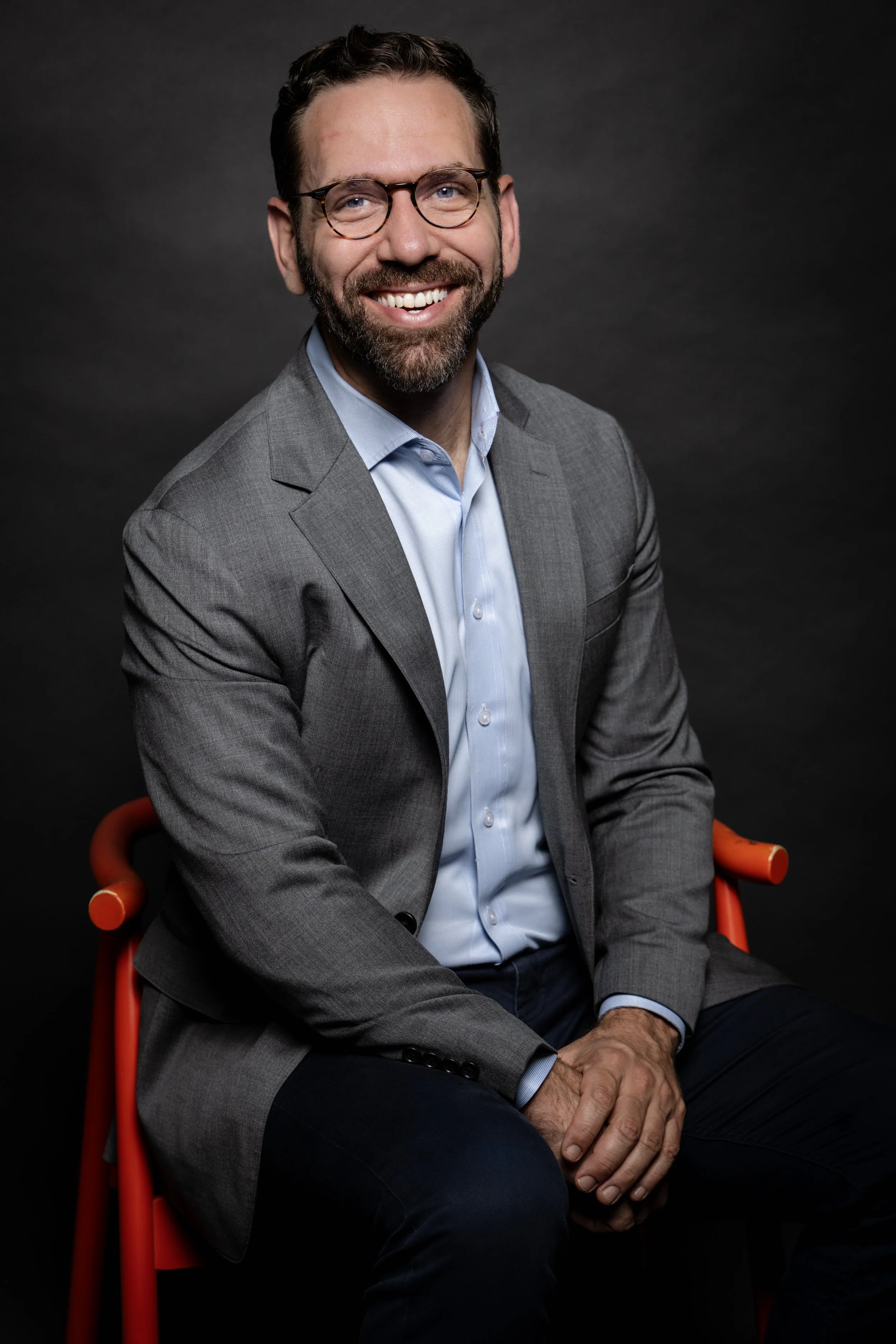 A man with a beard and glasses wearing a gray suit and light blue shirt, sitting on an orange chair, smiling against a dark background for a corporate headshot done in a studio