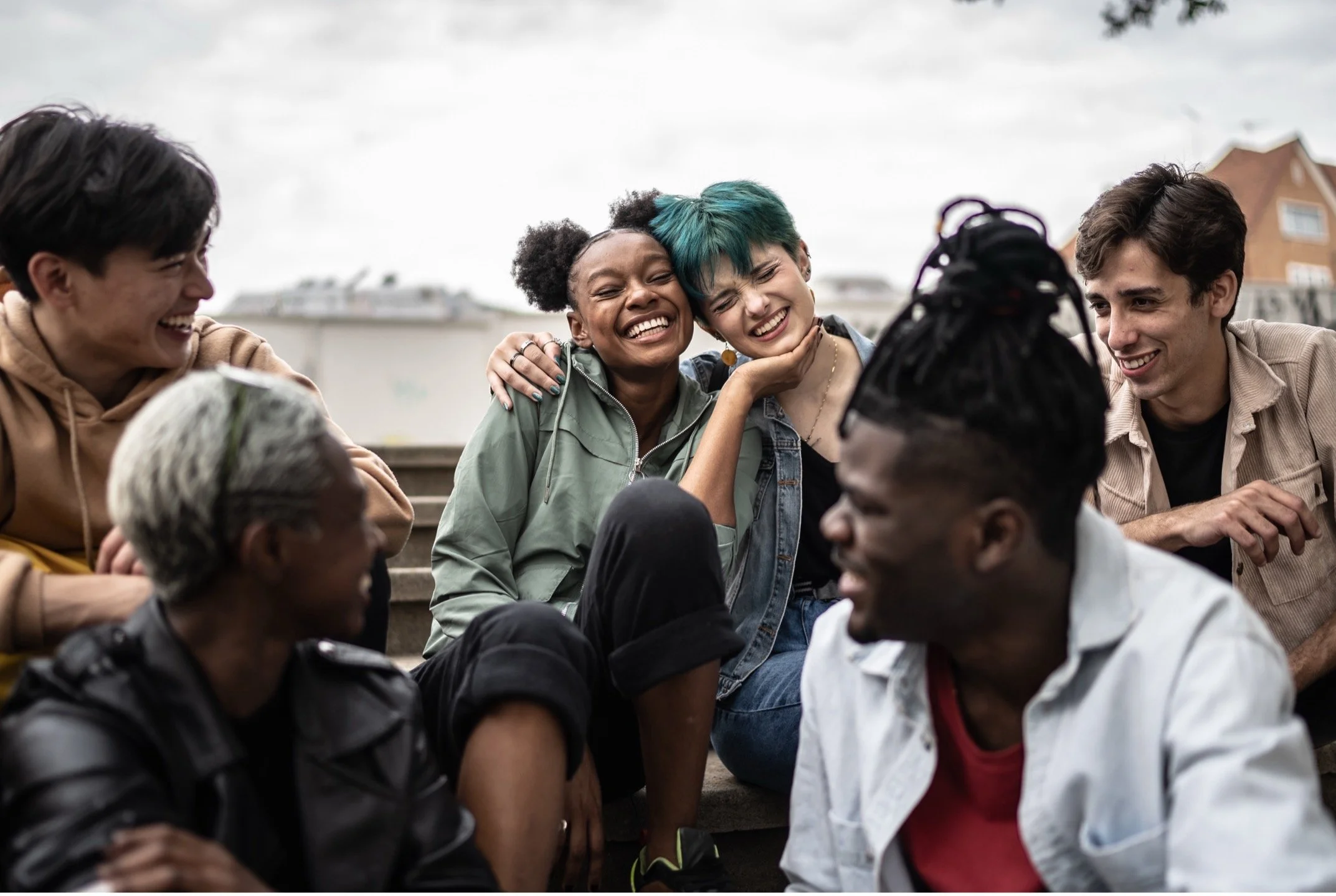 A diverse group of six young friends sitting outdoors on steps, smiling, laughing, and embracing each other, with a blurred urban background.
