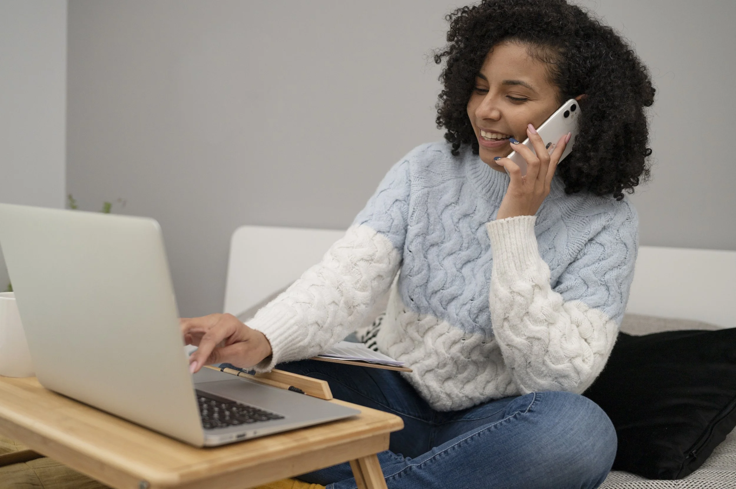 Woman with curly hair wearing a cable-knit sweater, sitting at a wooden table in front of a laptop, holding a smartphone to her ear, smiling, and looking at her laptop.