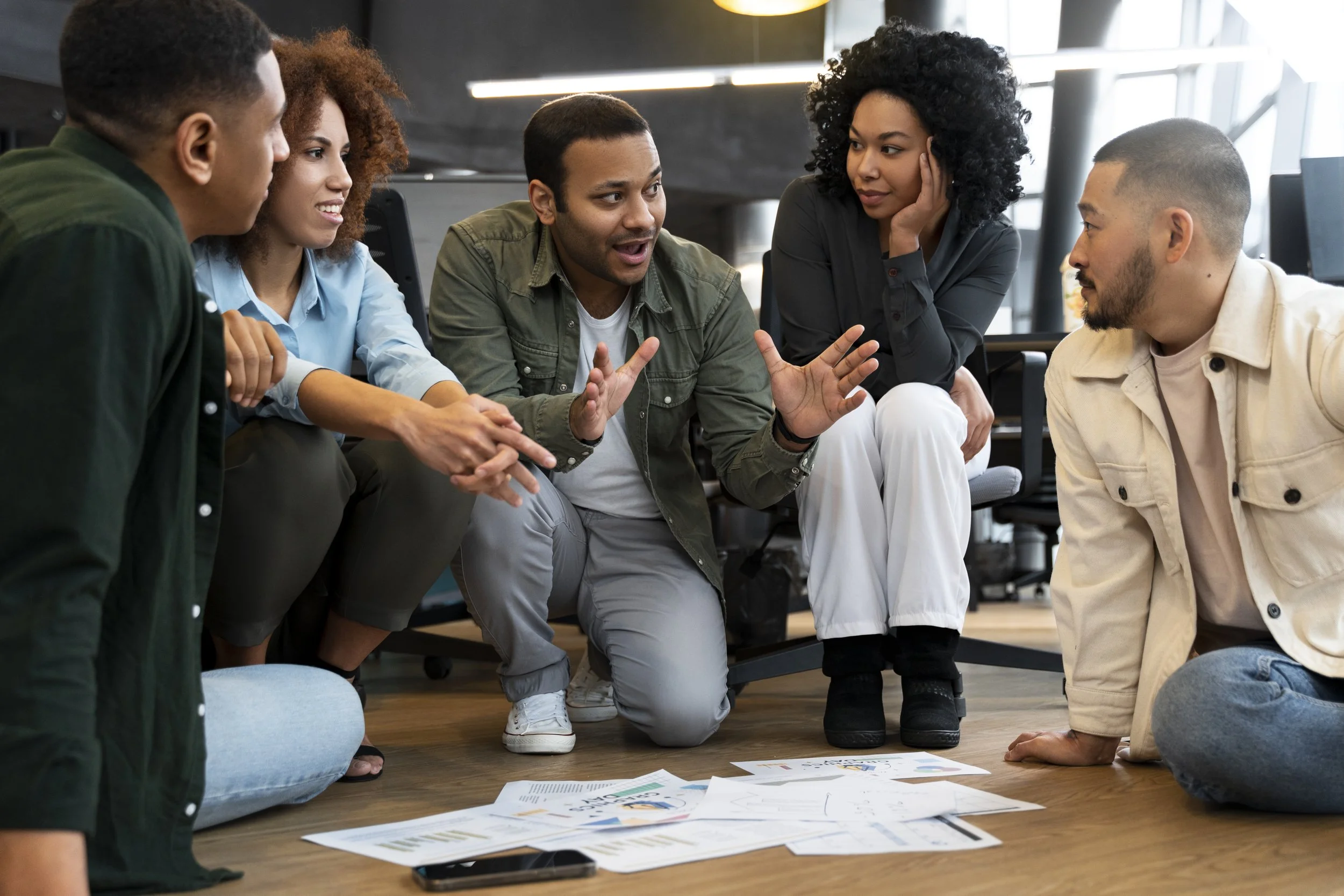 Group of five diverse young adults engaging in a discussion on the floor of an office, with papers scattered around them.
