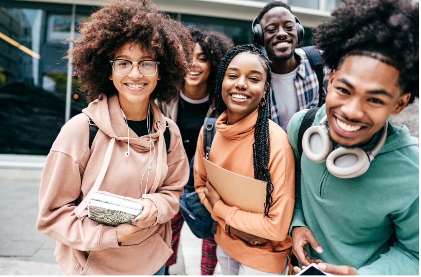 Five smiling young people standing together outdoors, holding books and wearing backpacks.