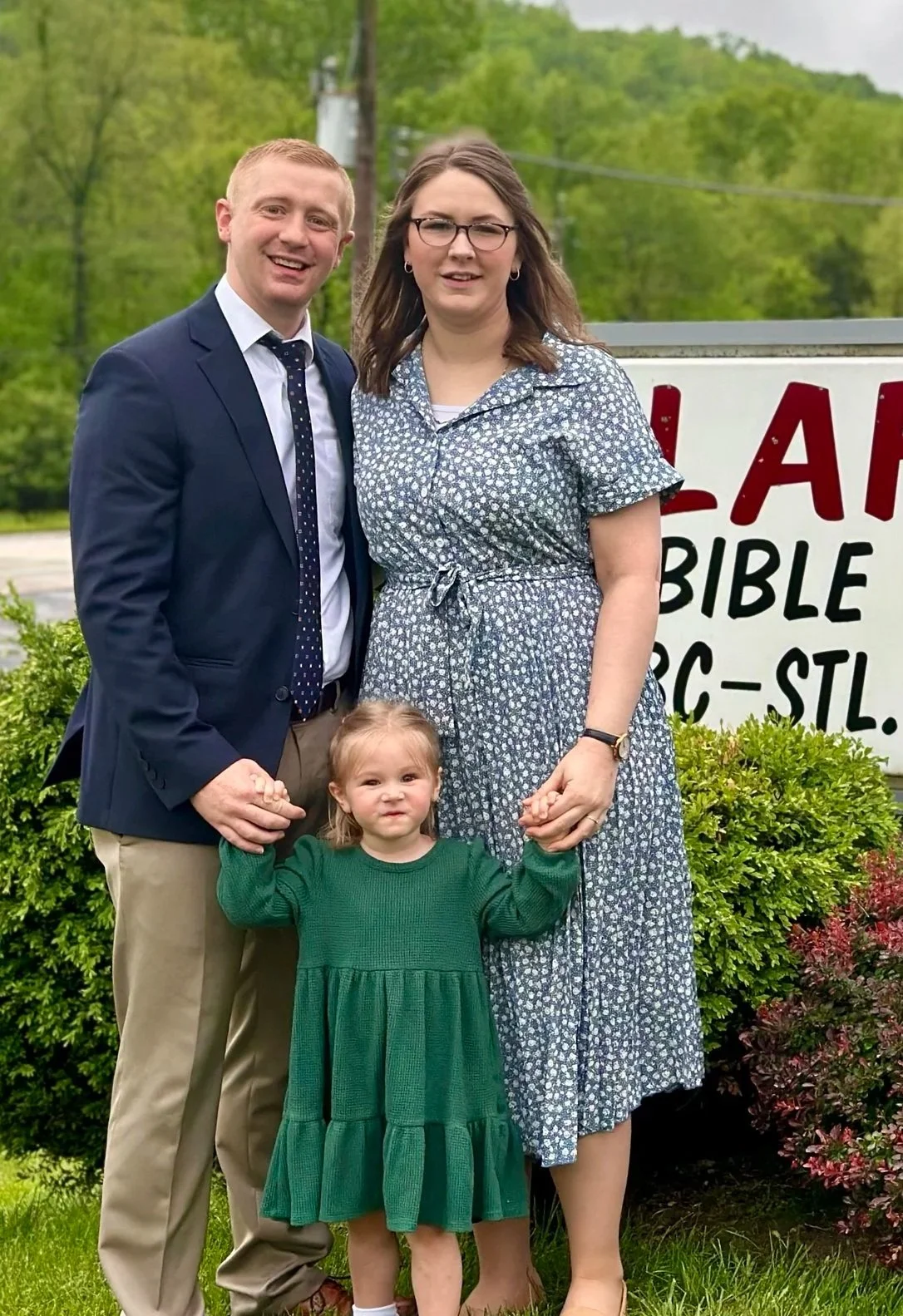 A family of three standing outdoors in front of a sign with trees in the background. The man is wearing a navy suit jacket, white shirt, and polka dot tie, holding the hand of a young girl in a green dress. The woman is wearing glasses and a blue floral dress, holding the child's other hand. The little girl has a serious expression and is dressed in a green dress.