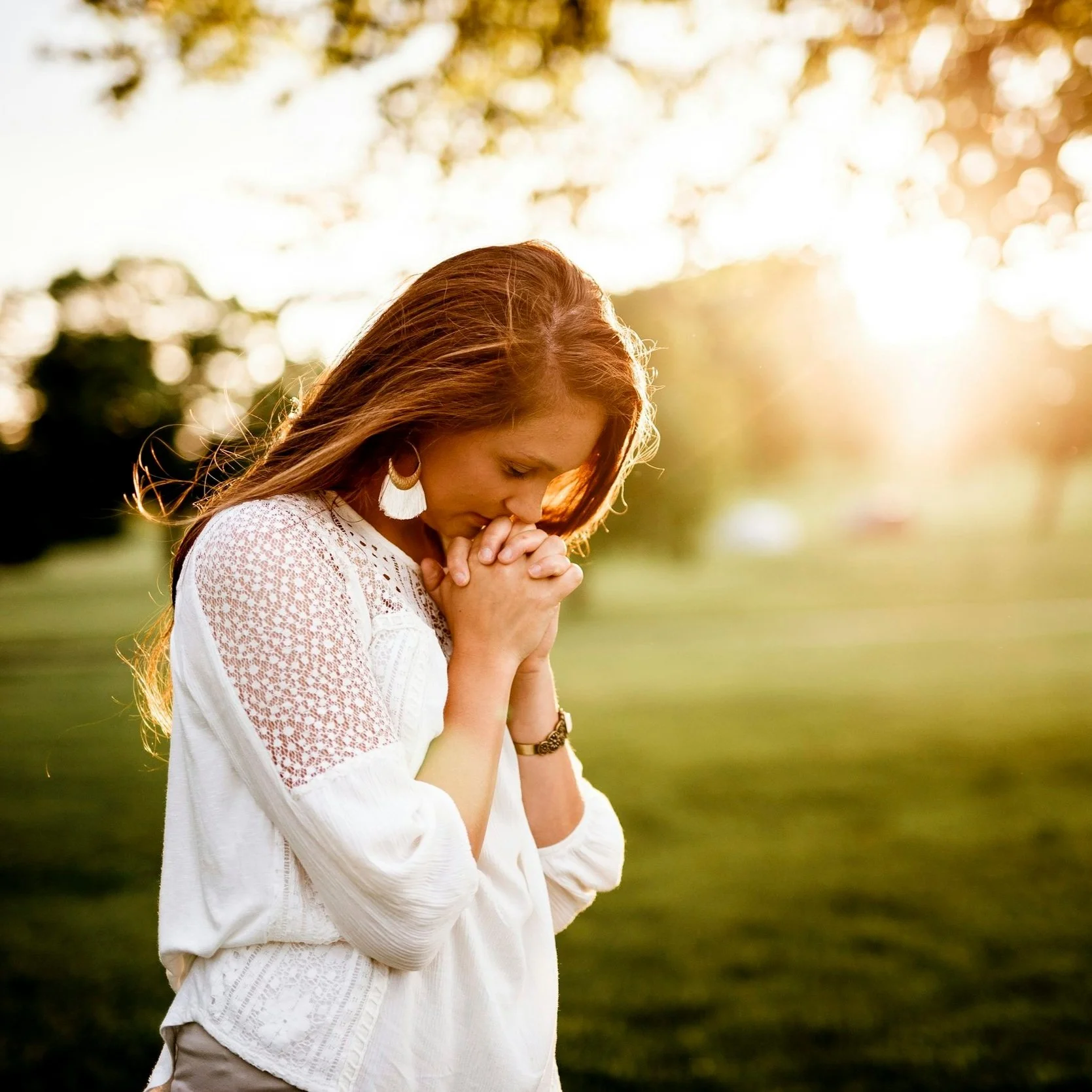A woman with long red hair standing outdoors with her hands clasped in prayer, eyes closed, during sunset, wearing a white shirt and earrings.