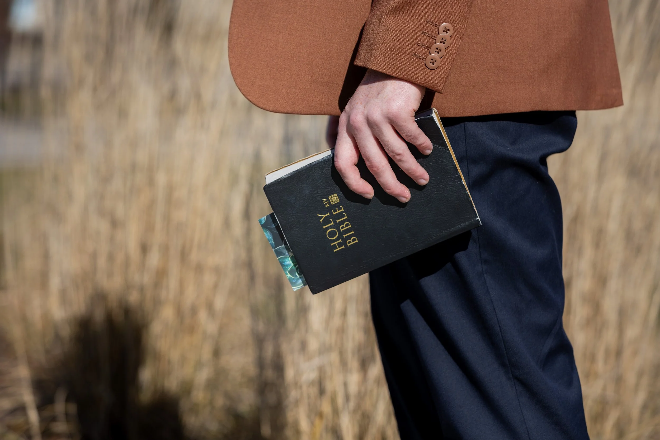 Person in a brown jacket holding a black Holy Bible with a colorful bookmark, standing outdoors on dry grass.