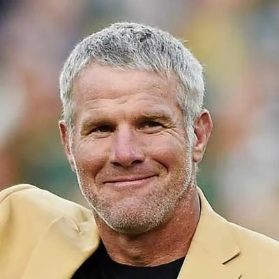 Brett Farve with short gray hair, smiling, wearing a beige blazer, outdoors with blurred colorful background.