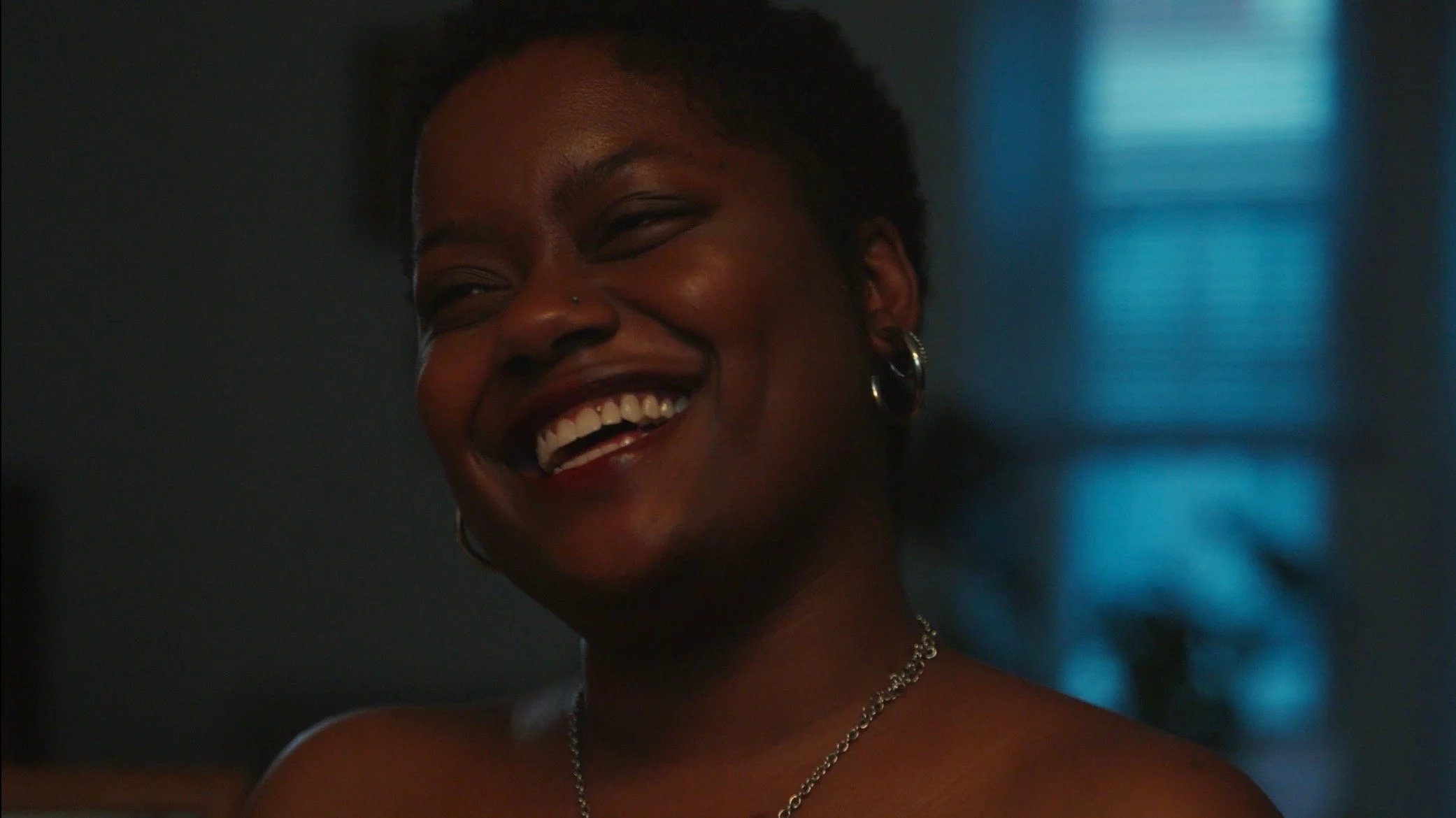 A woman with dark skin and short curly hair smiling joyfully, wearing earrings and a necklace, indoors with blue light in the background.