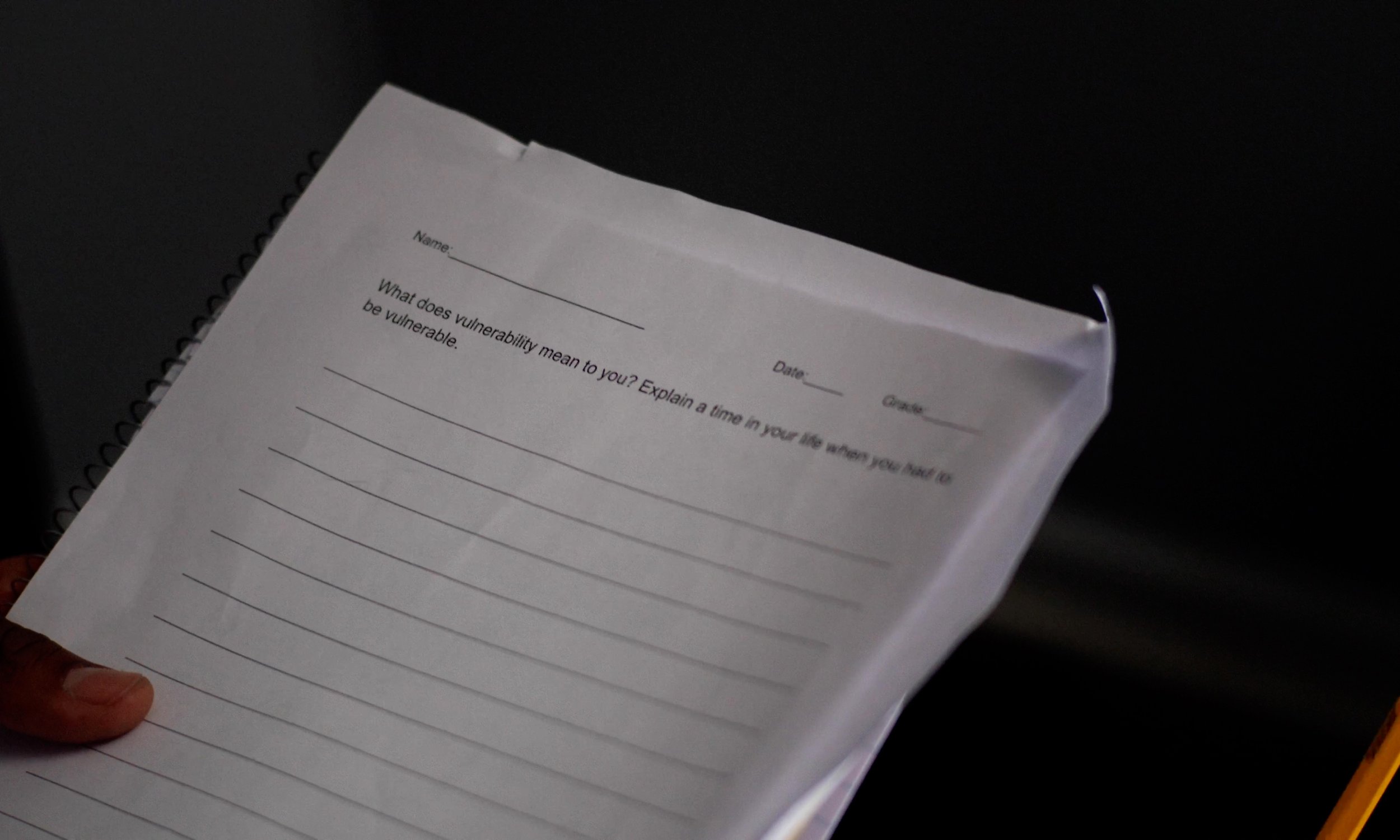 A hand holding a spiral-bound notebook with a worksheet titled 'What does vulnerability mean to you? Explain a time in your life when you had to be vulnerable.'