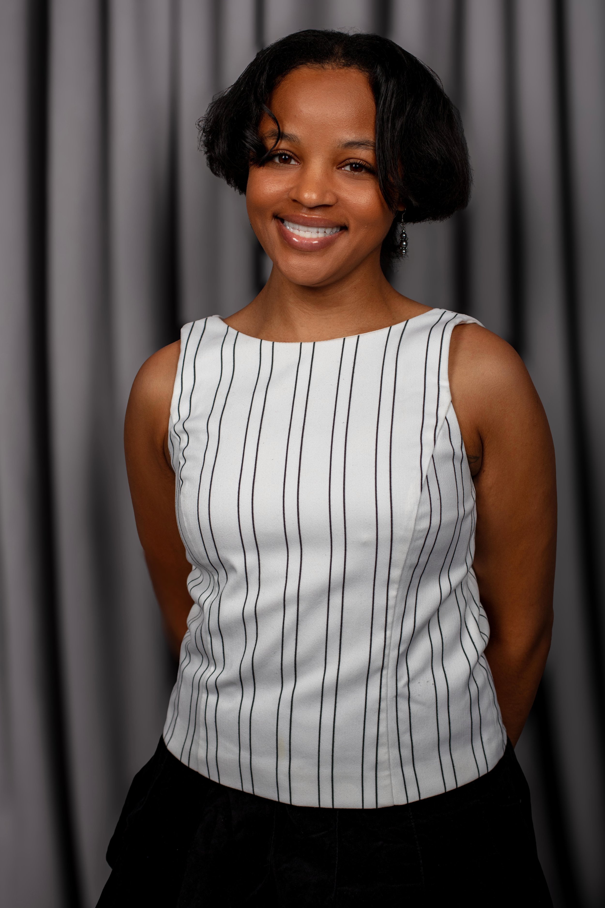 A woman with short black hair wearing a white sleeveless top with black vertical stripes, smiling in front of a grey curtain background.