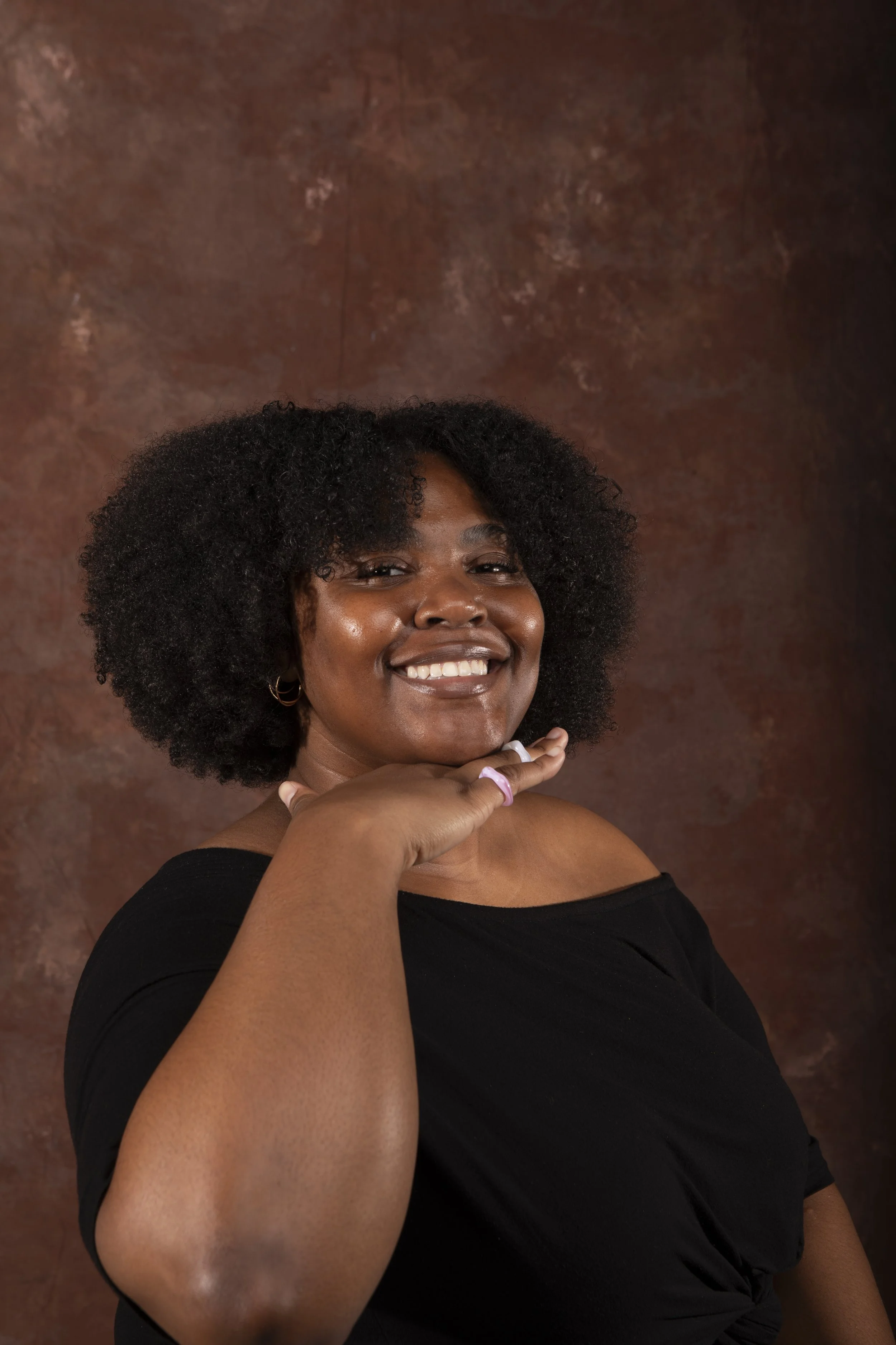 A smiling woman with dark skin and curly hair, wearing a black top, touching her chin with her hand, standing against a brown textured background.