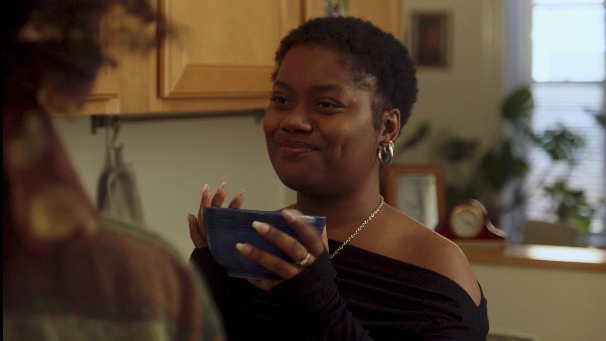 A woman with short curly hair and jewelry smiling while holding a blue bowl indoors.