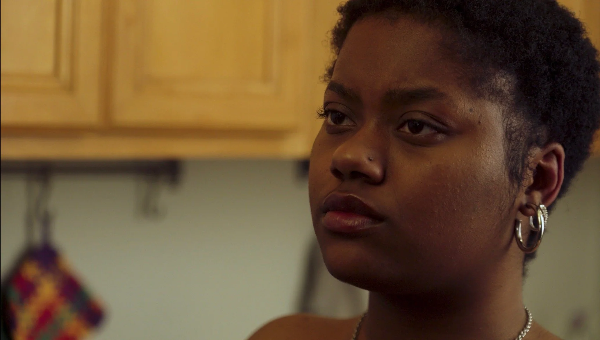 A young Black woman with short hair and earrings, looking to the side in a kitchen with wooden cabinets