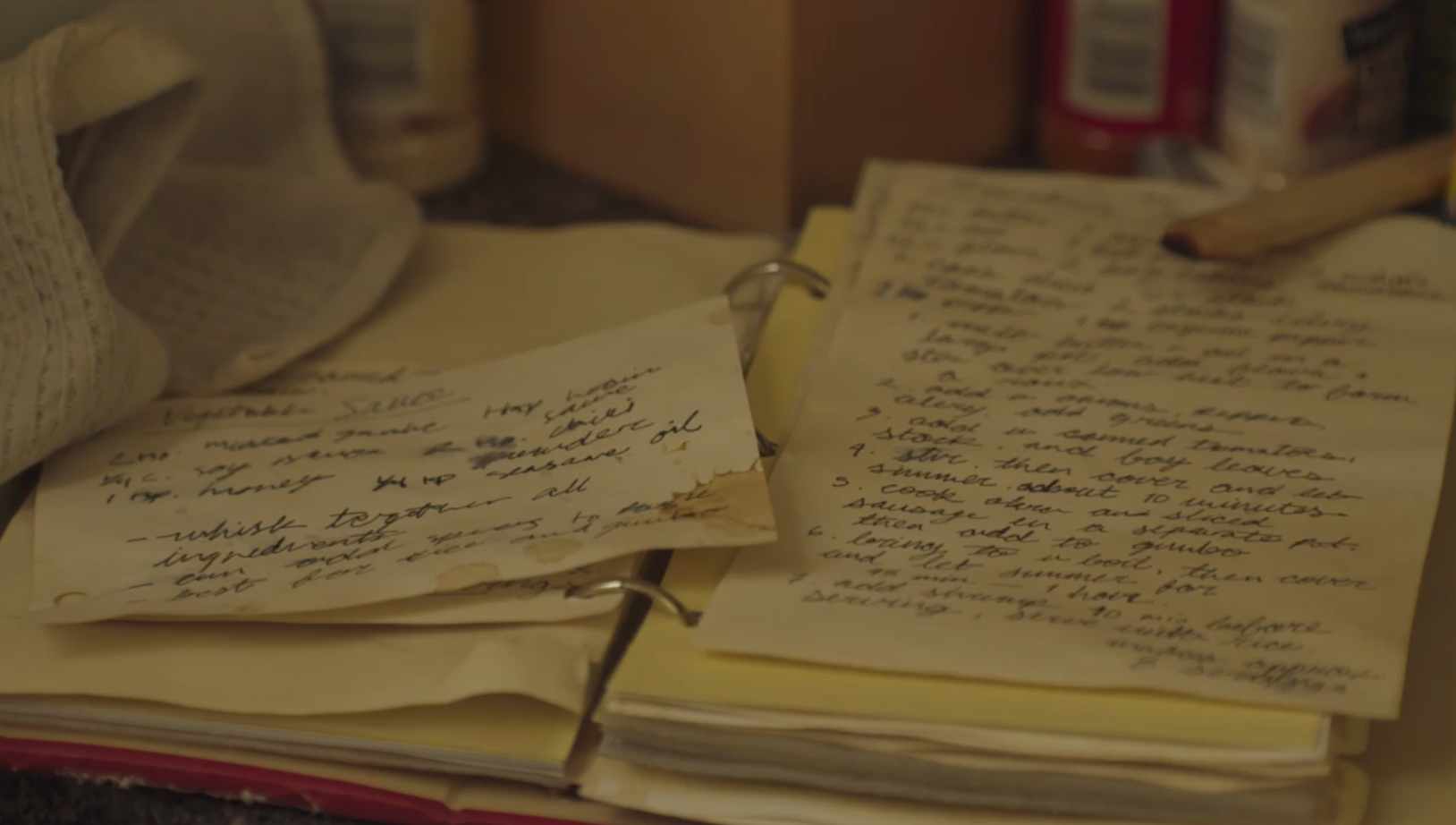 Handwritten recipes on scrap paper and a binder, surrounded by grocery items and a tea towel.