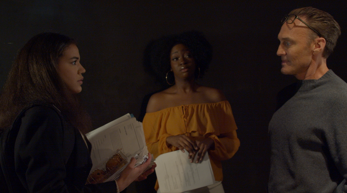 Three people in conversation, one woman holding documents, one woman in yellow off-shoulder top, another man in glasses and gray sweater, dark background.