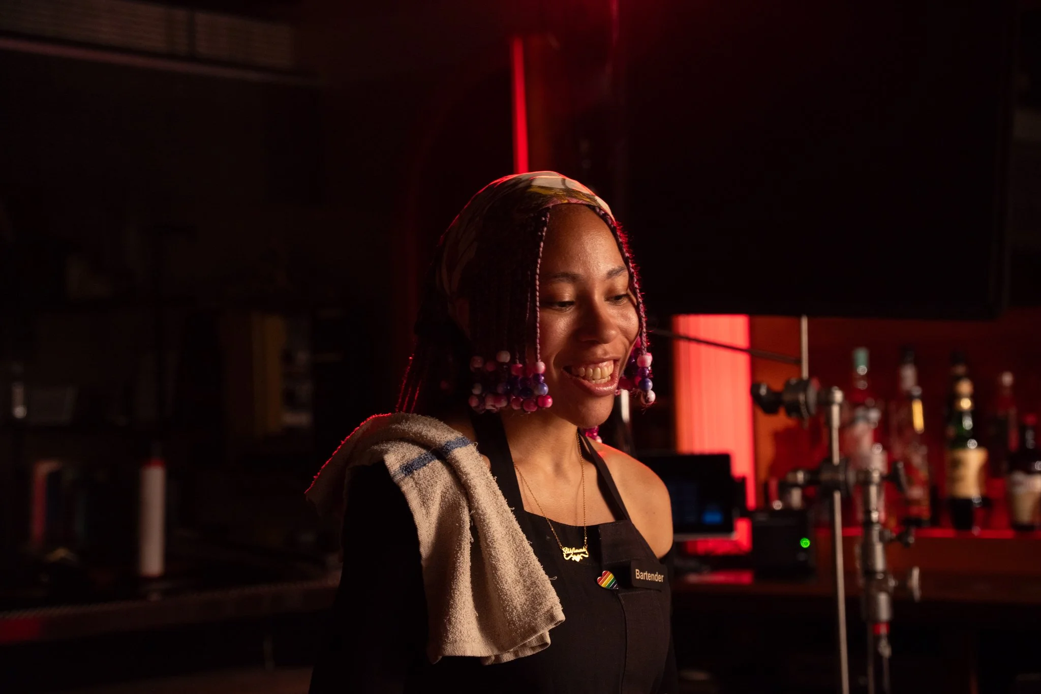 A smiling bartender with colorful hair beads and a towel over her shoulder, standing in a dimly lit bar with bottles and equipment in the background.