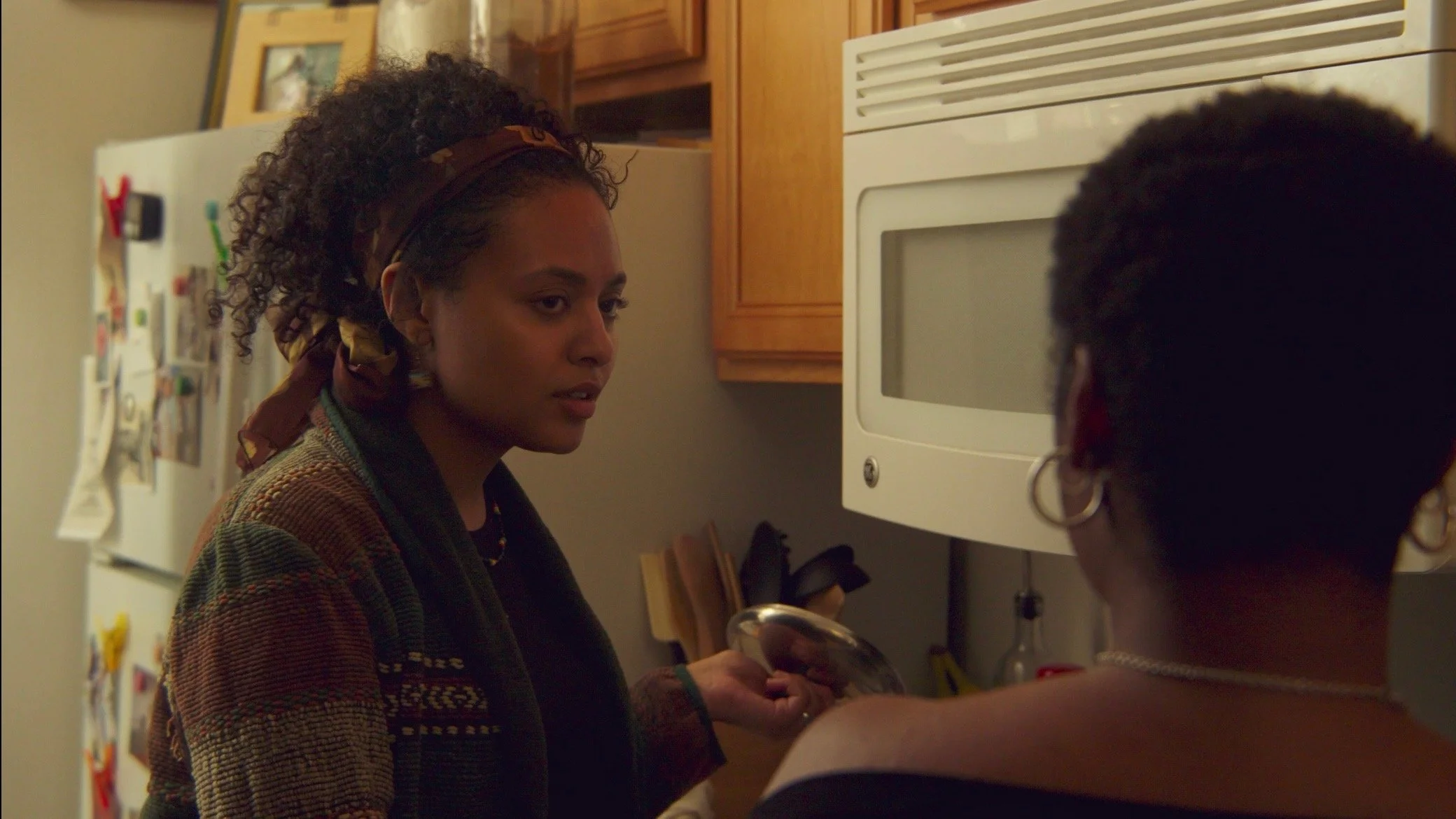 Two women having a serious conversation in a kitchen, one holding a dish, with a refrigerator and microwave in the background.