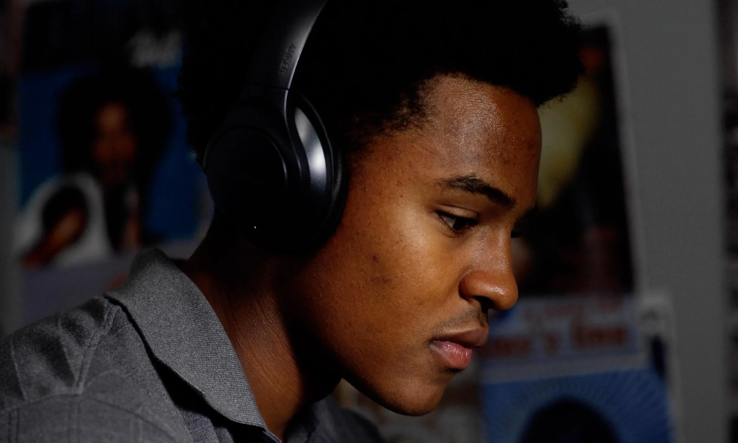 A young man with dark skin and short curly hair wearing black over-ear headphones and a gray shirt, focused on using a computer.