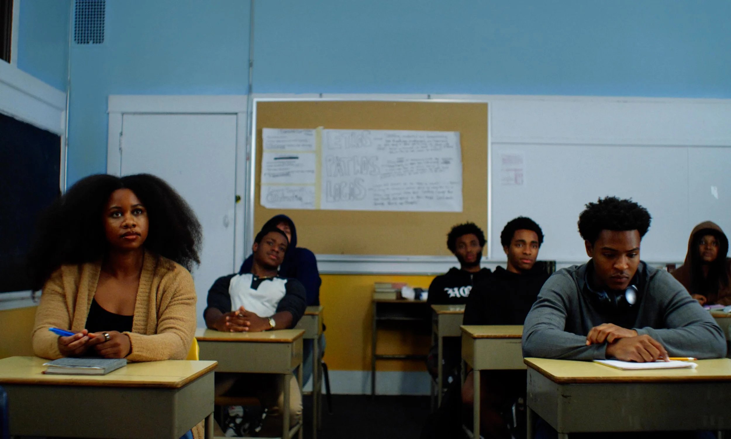 Classroom scene with students sitting at desks, paying attention; some with notebooks and pens, a blackboard, and a bulletin board in the background.