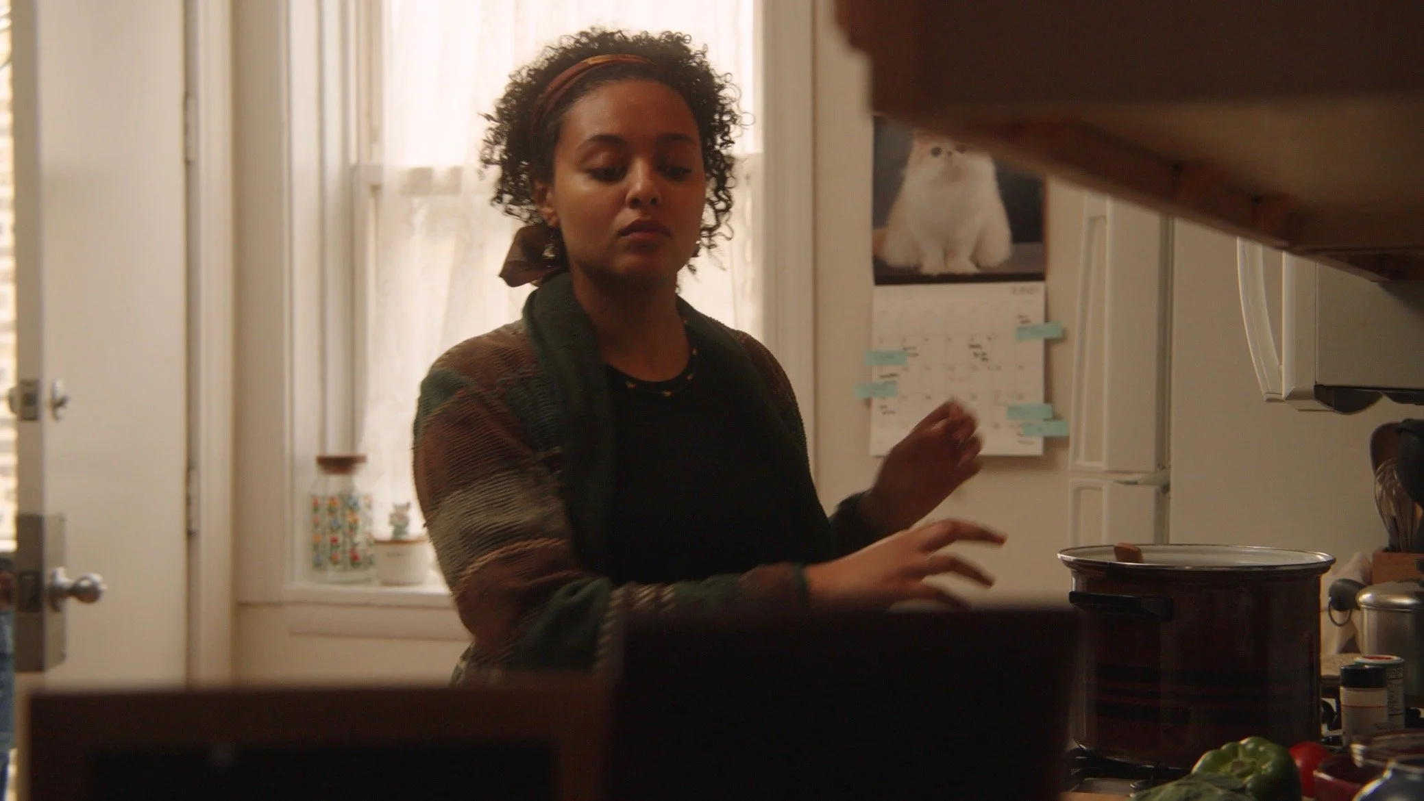 A woman with curly hair and a headband stands in a kitchen near a window, looking down and reaching towards a slow cooker on the counter.
