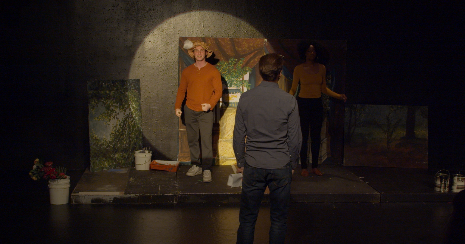 Three people on a small stage practicing acting, with painted backdrops and props around them.