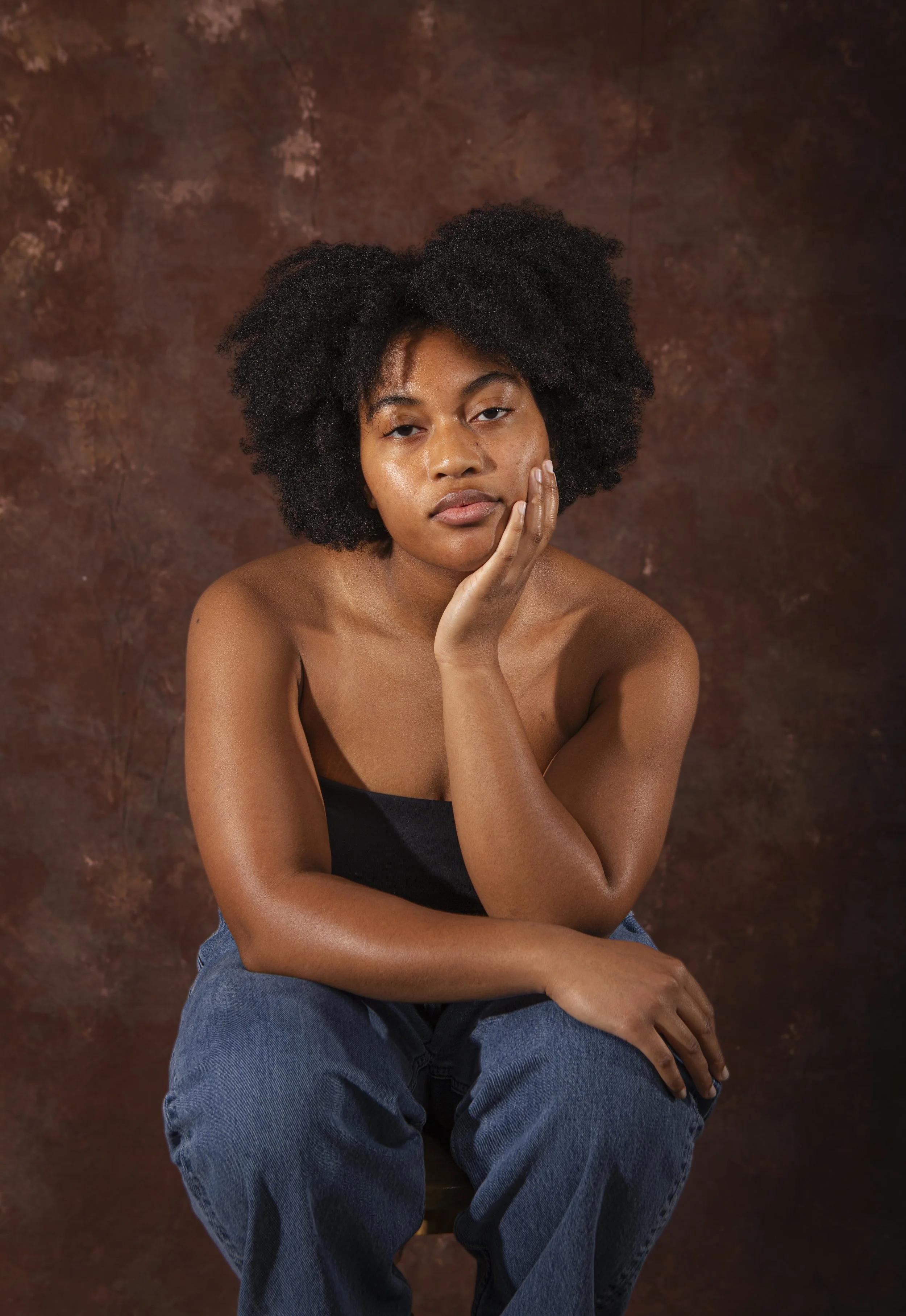 A young woman with natural curly hair, sitting against a brown textured background, wearing a black strapless top and blue jeans, resting her chin on her hand.