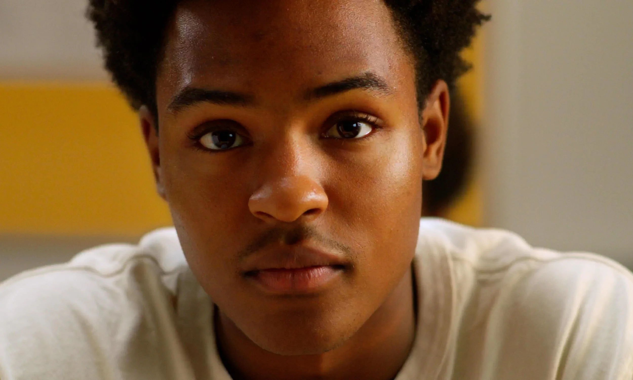 Close-up of a young man with short, curly black hair, wearing a white shirt, looking directly at the camera.