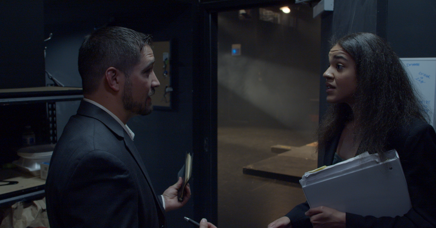 Man and woman having a serious conversation in an industrial or storage area, with the woman holding a clipboard and papers, and the man holding a notepad.