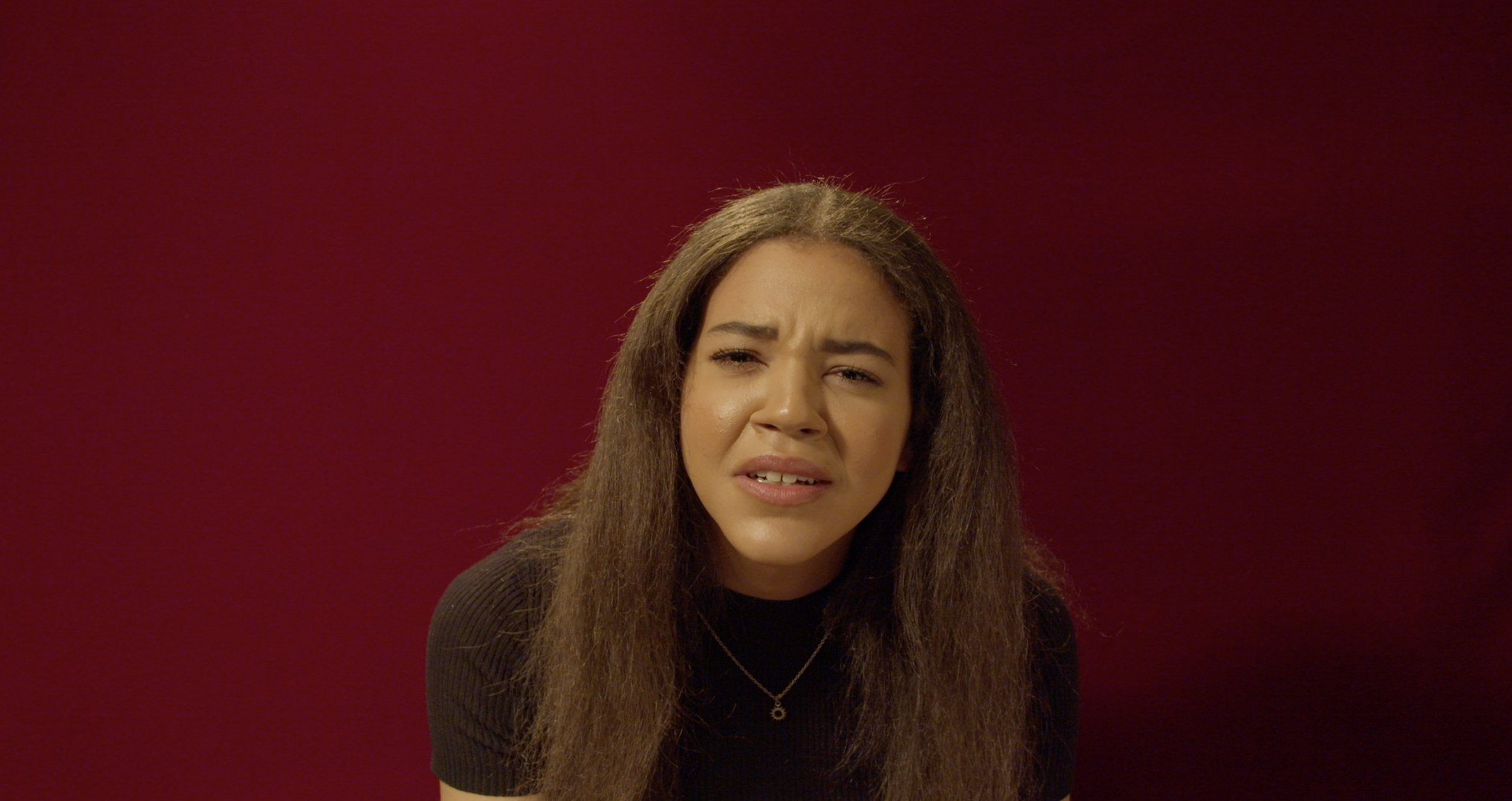 A young woman with long brown hair and a puzzled or confused expression, wearing a black top and a small necklace, against a plain red background.