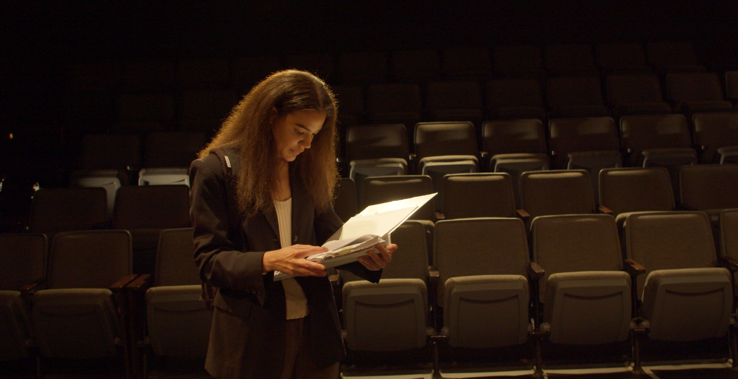A woman with long, curly hair standing in an empty theater or auditorium, reading a script or document illuminated by a light.