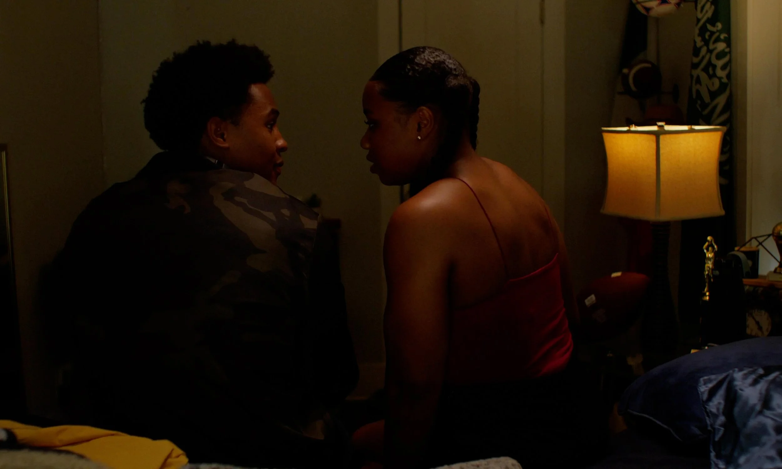 A man and woman sitting closely on a bed in a dimly lit room, facing each other and engaging in a conversation.