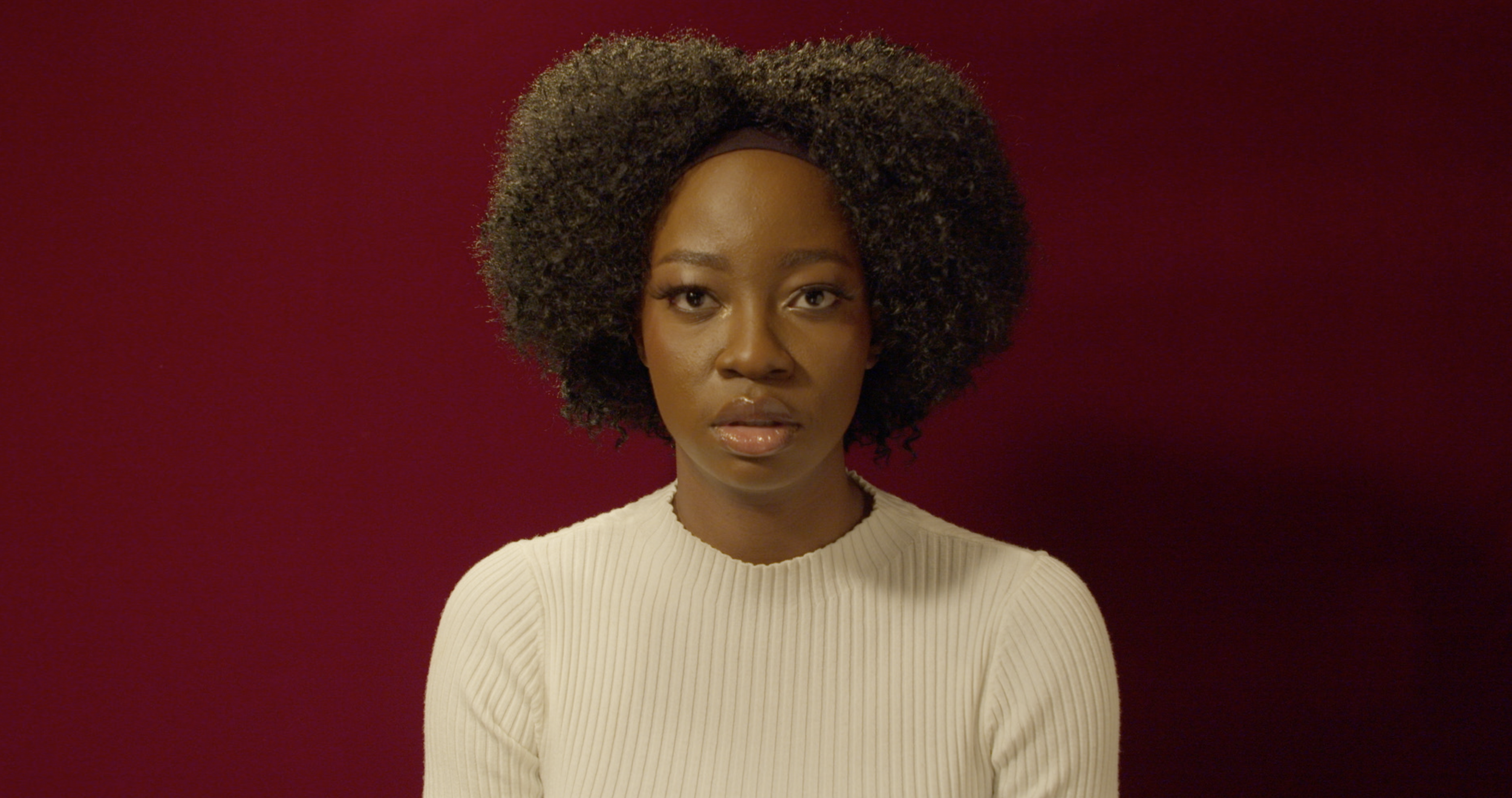 A woman with natural curly hair wearing a cream-colored ribbed sweater, standing against a red background, looking directly at the camera with a serious expression.