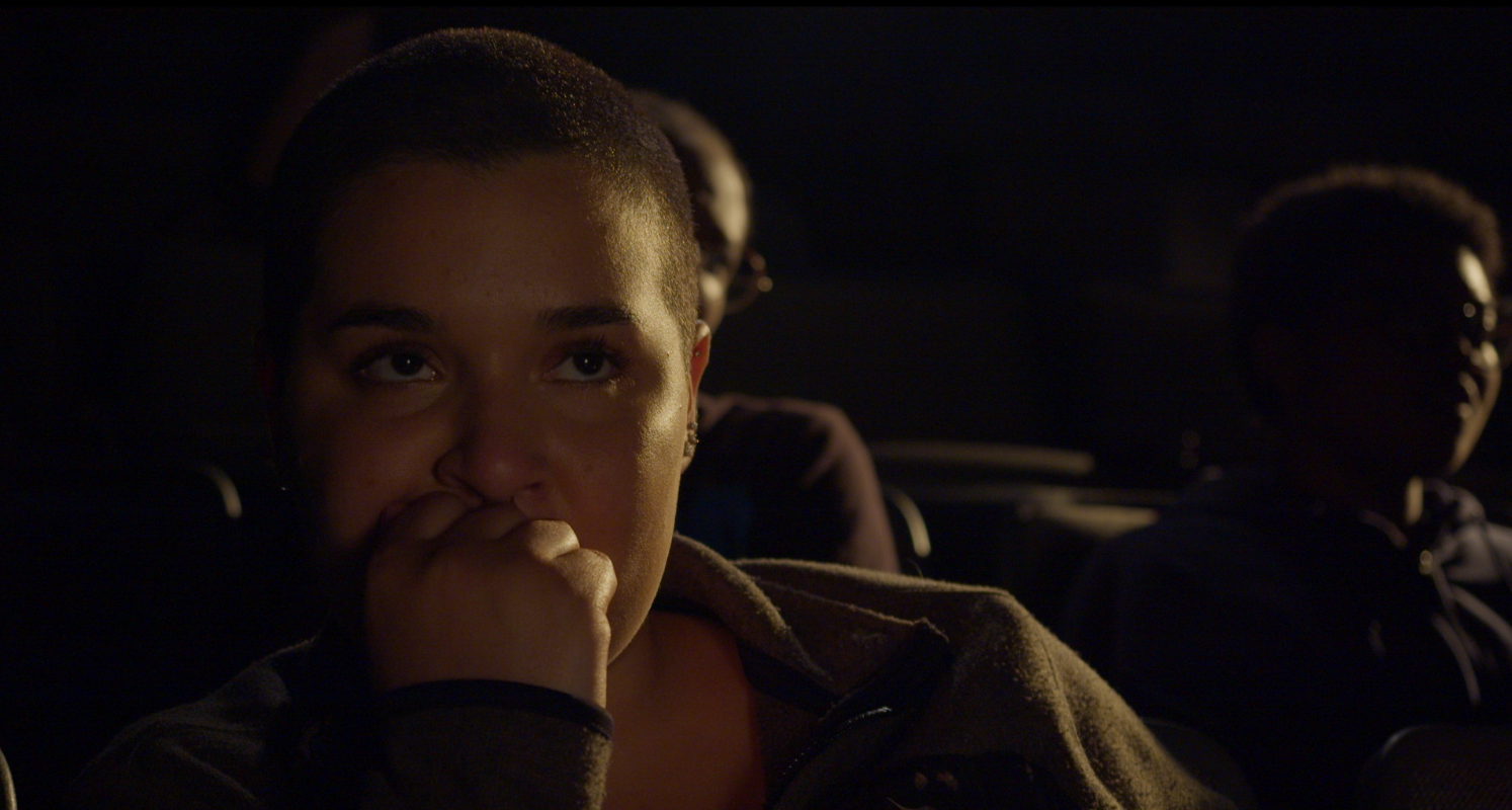 A young woman with short hair looks intensely at the camera, resting her chin on her hand, sitting in a dark theater or auditorium with other people visible in the background.
