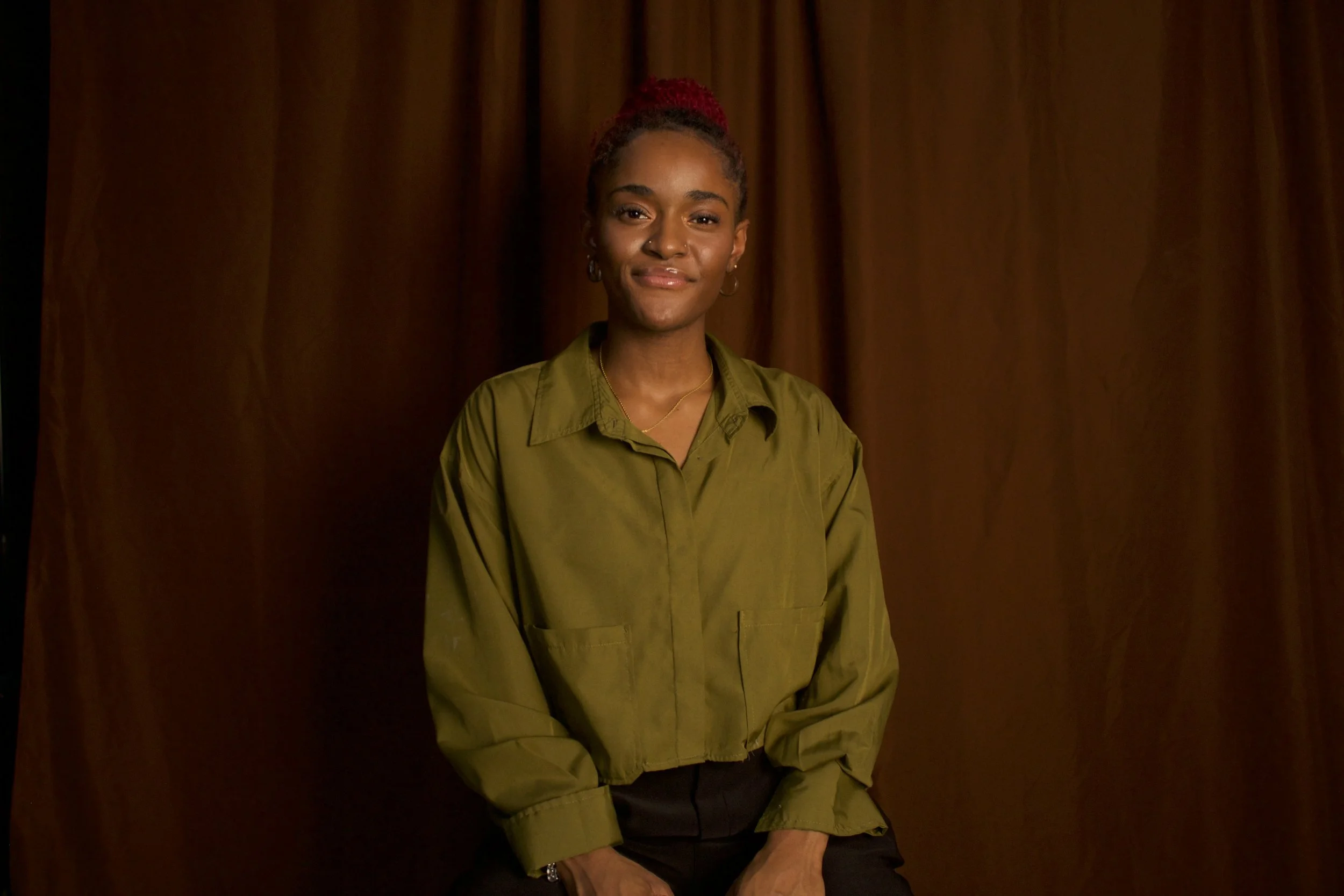 A young woman with red hair pulled back, wearing an olive green jacket, sitting in front of a brown curtain backdrop, looking at the camera with a slight smile.