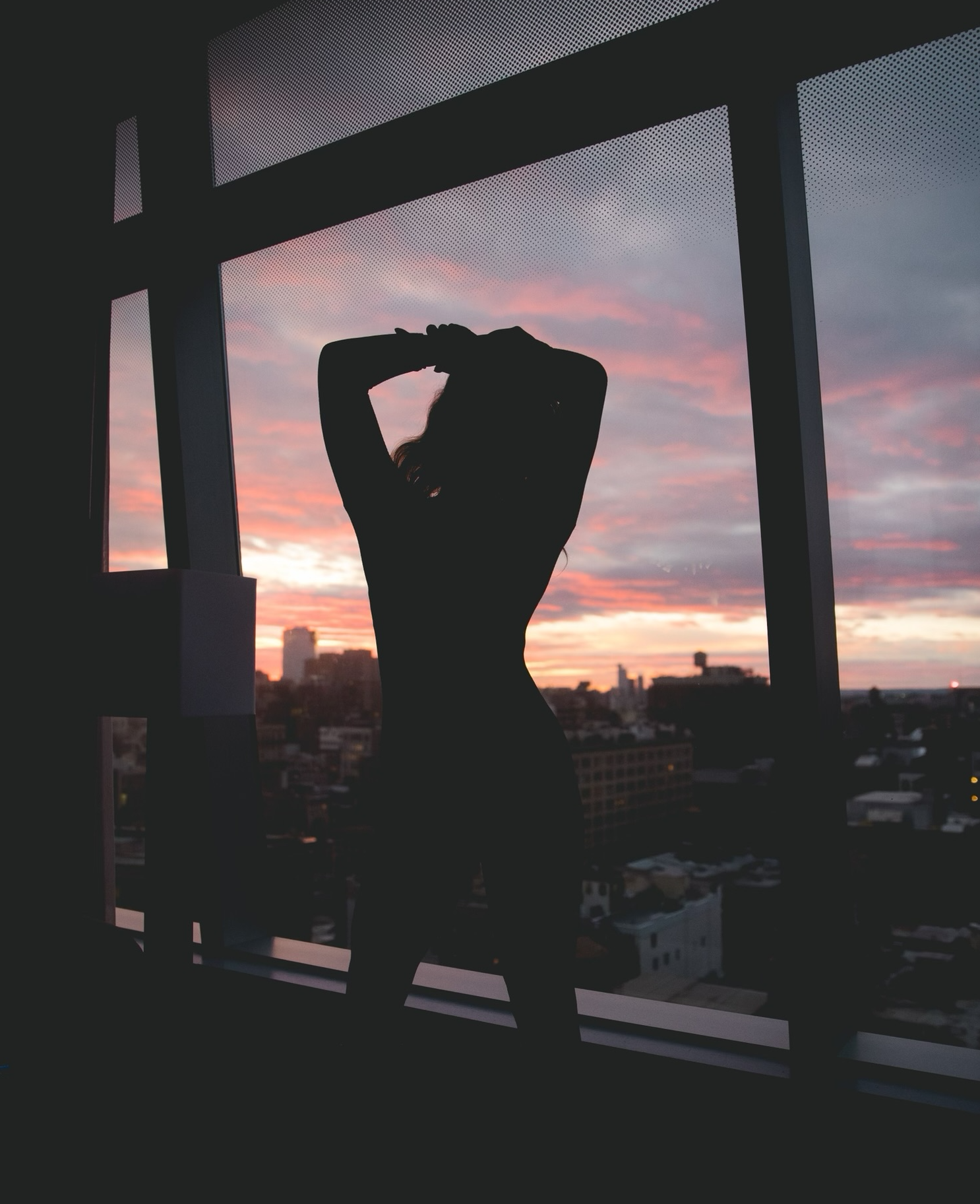 Silhouette of a person standing by a large window with a sunset view over a cityscape.
