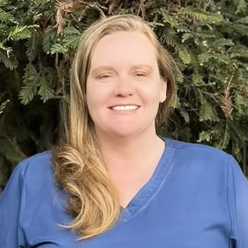 A woman with long, wavy blonde hair wearing a blue shirt, smiling in front of green foliage.