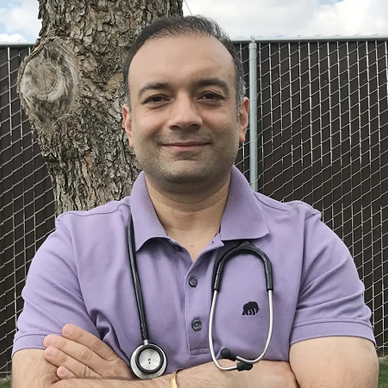 A man standing outdoors with arms crossed, wearing a purple polo shirt and a stethoscope around his neck, in front of a tree and a chain-link fence.
