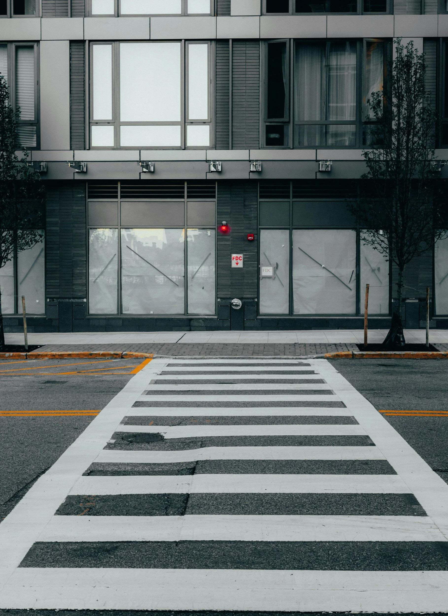 Urban crosswalk in front of modern building with large windows, trees, and fire department connection signs.