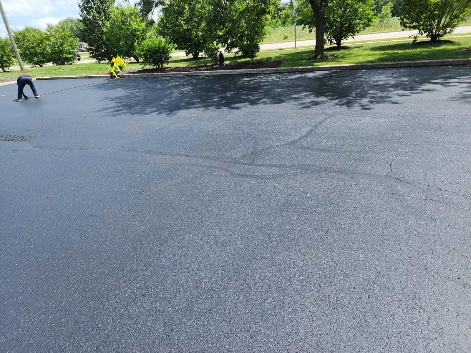 Freshly paved asphalt parking lot with two people working, surrounded by trees and grass.
