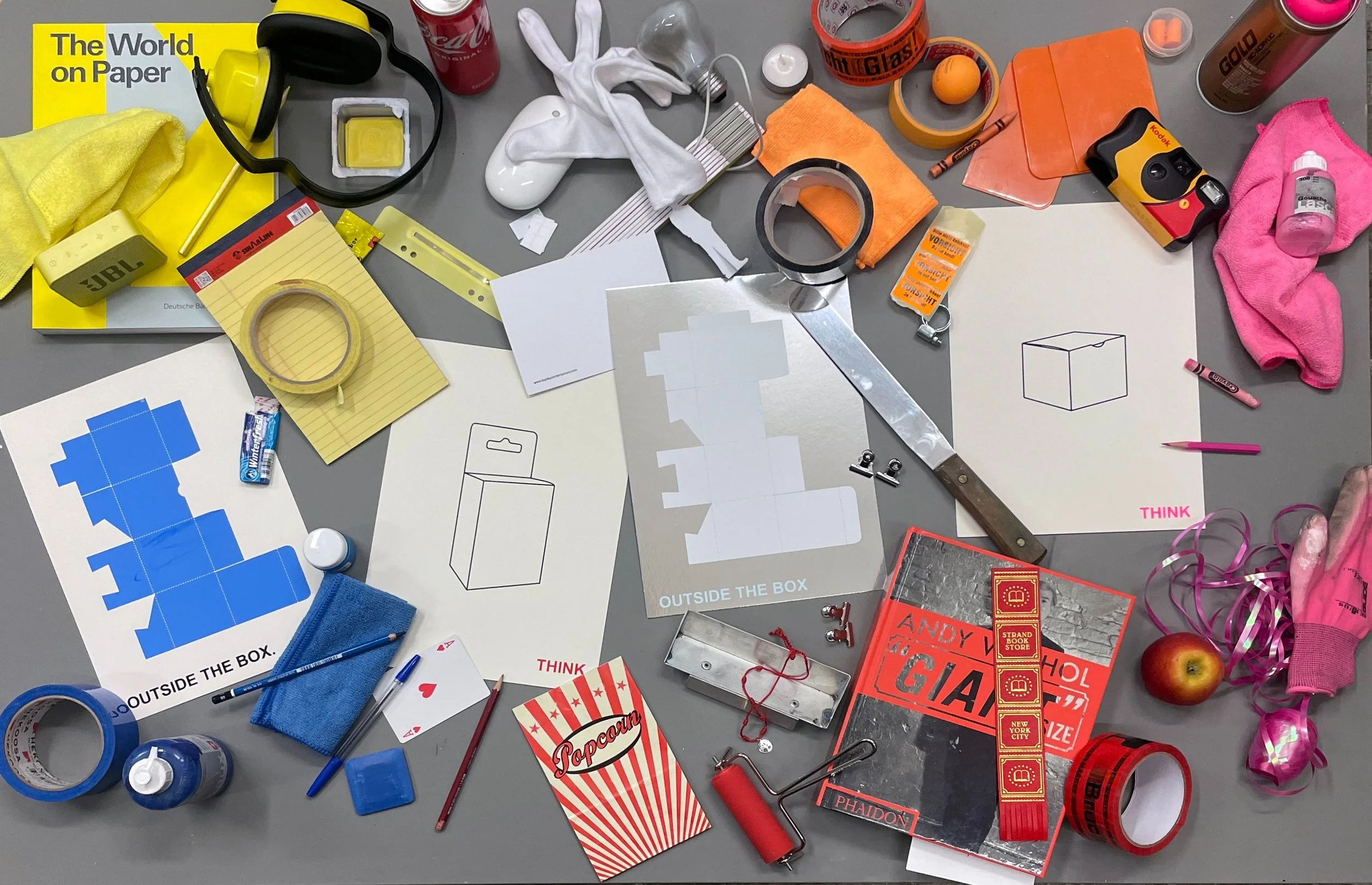 A cluttered table filled with various objects from the screen printing studio with the think print surrounded by scissors, markers, spray bottles, a glove, an apple, a toy lig,  supplies, suggesting a workspace for a creative or project task.