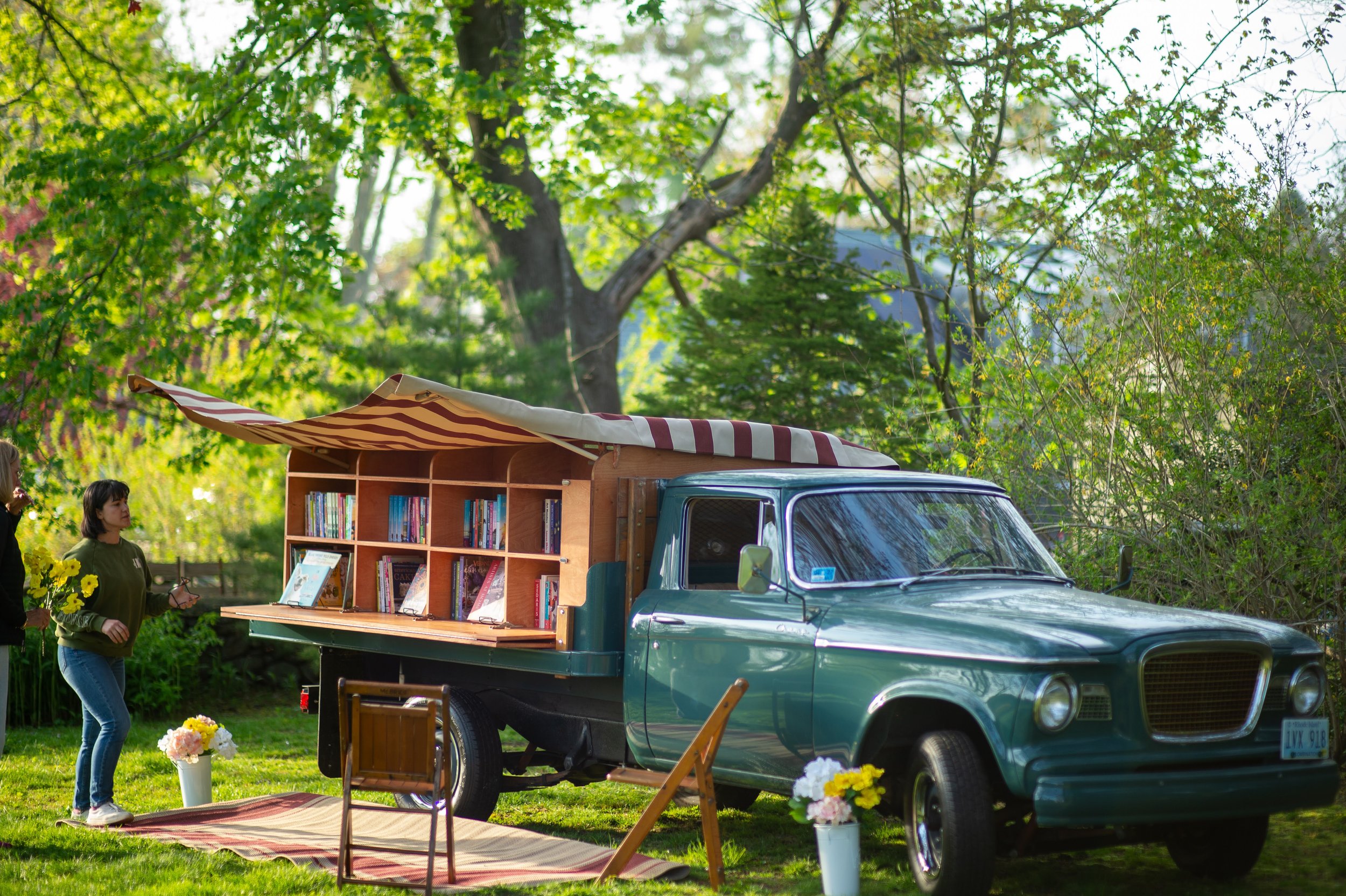 A vintage green pickup truck parked in a grassy area, converted into a mobile bookshop with an awning displaying shelves of books. Two people stand nearby, and flowers and a small chair are arranged on the ground.