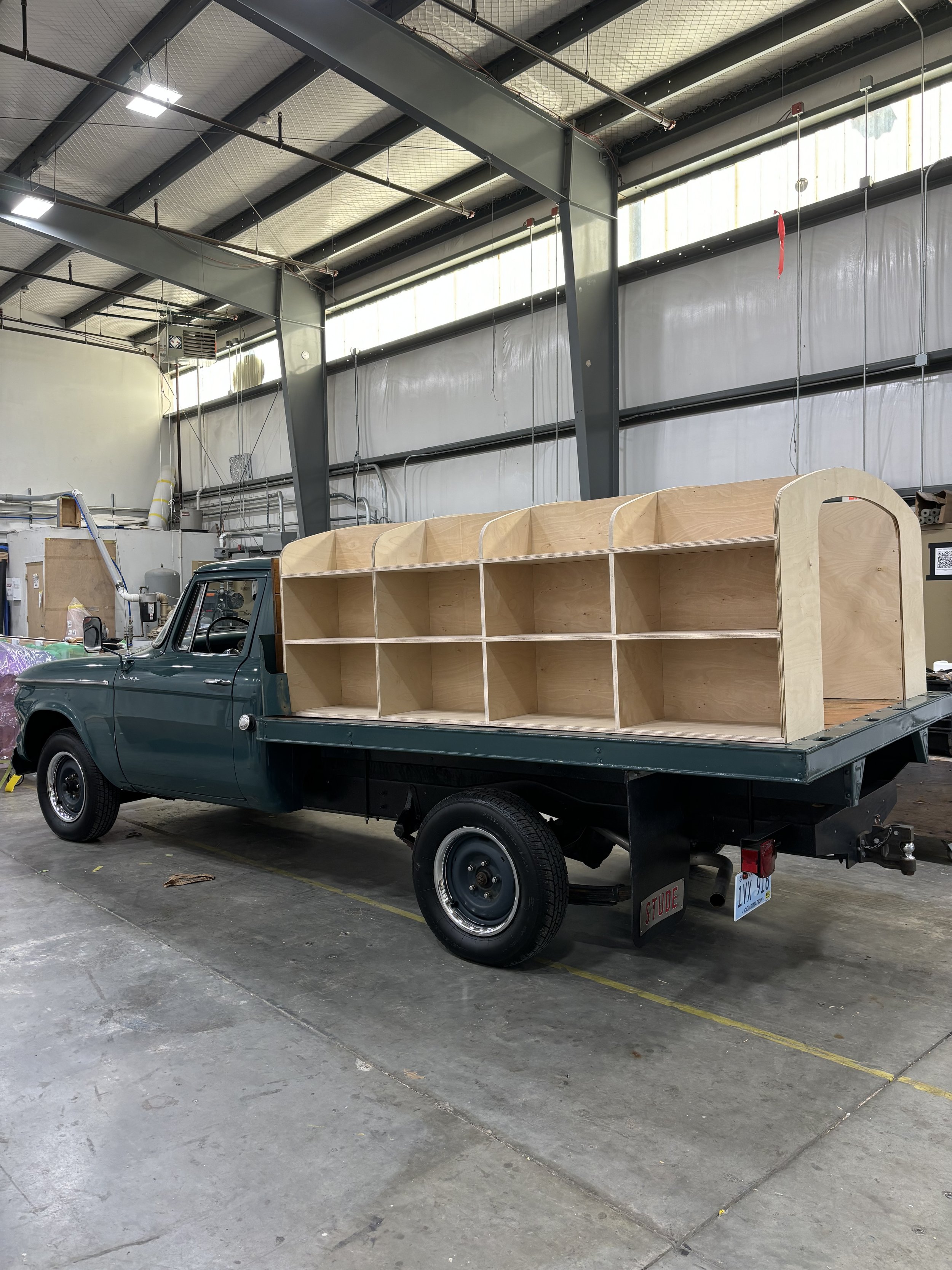 A vintage flatbed truck with wooden shelving installed on the bed, parked inside an industrial warehouse.