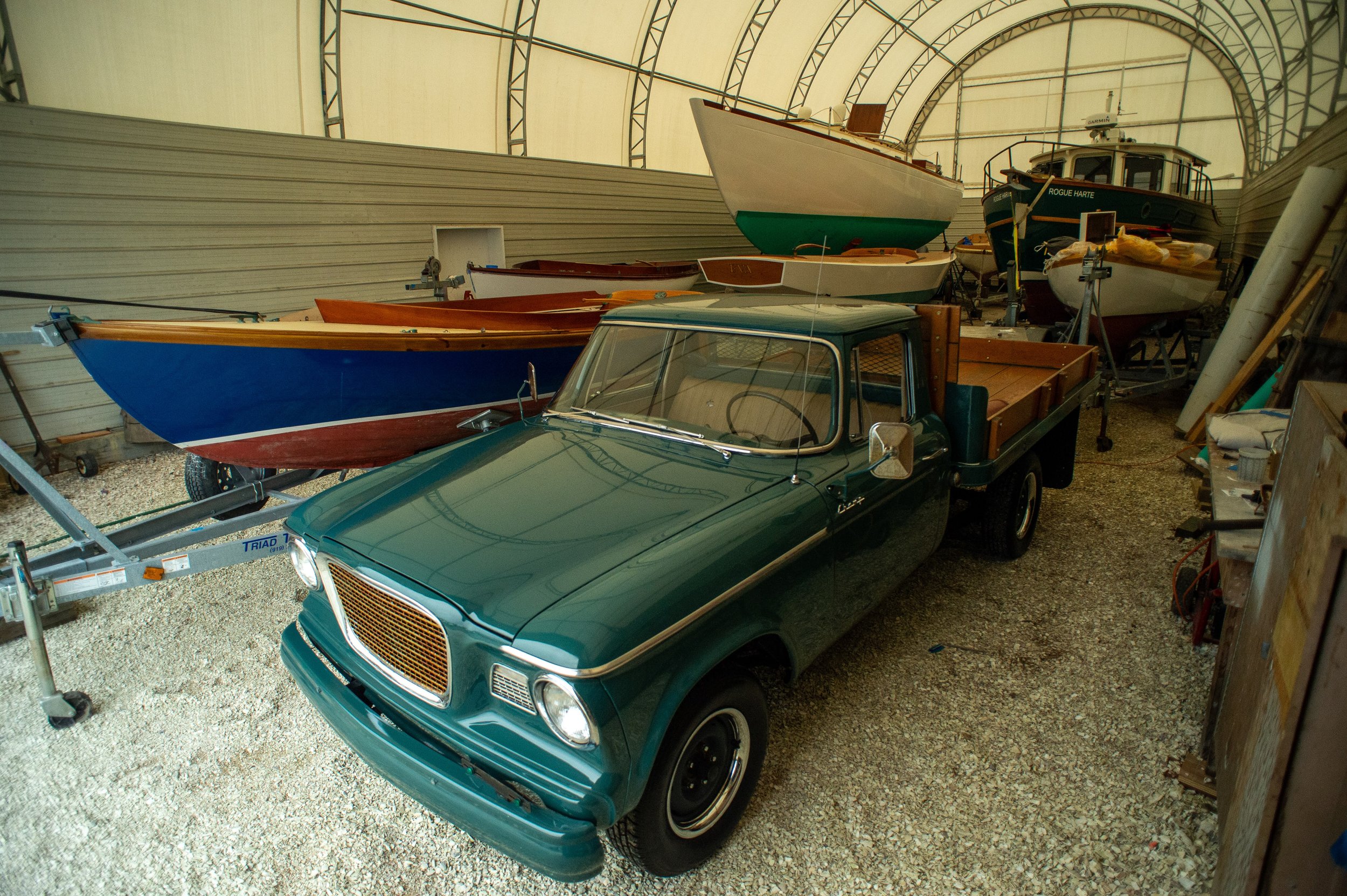 Green vintage truck parked inside a storage area with several boats on trailers, surrounded by tools and equipment.