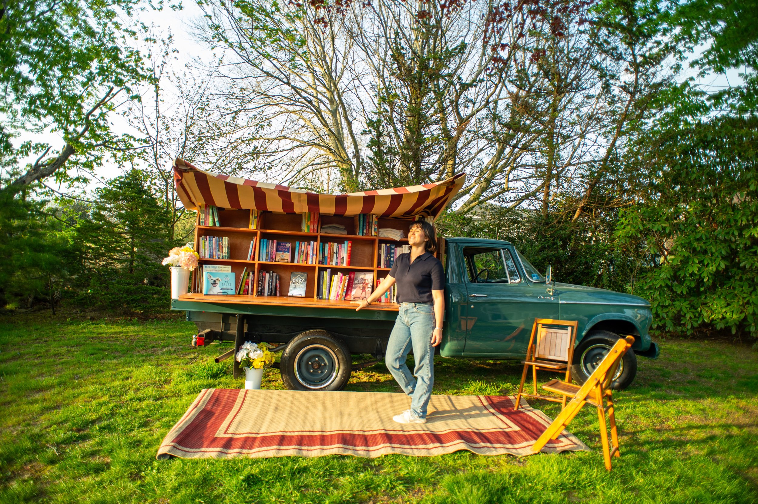 A person standing next to a green vintage truck converted into a mobile library, with bookshelves and a striped awning, parked on grass with surrounding trees.