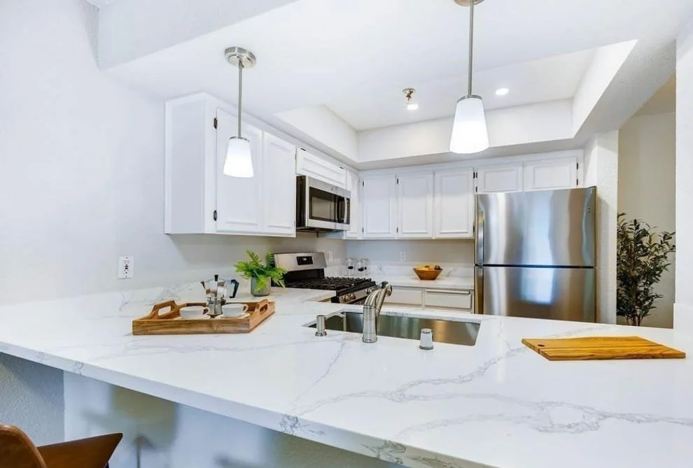 Modern kitchen with white cabinets, stainless steel refrigerator, microwave, black stove, and a marble countertop with a wooden cutting board and a tray with cups and a plant