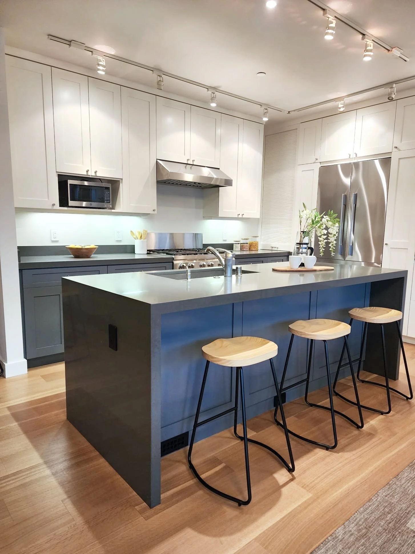 Modern kitchen with white upper cabinets and dark lower cabinets, stainless steel appliances, a kitchen island with a light gray countertop, three wooden bar stools with black metal legs, and hardwood flooring.