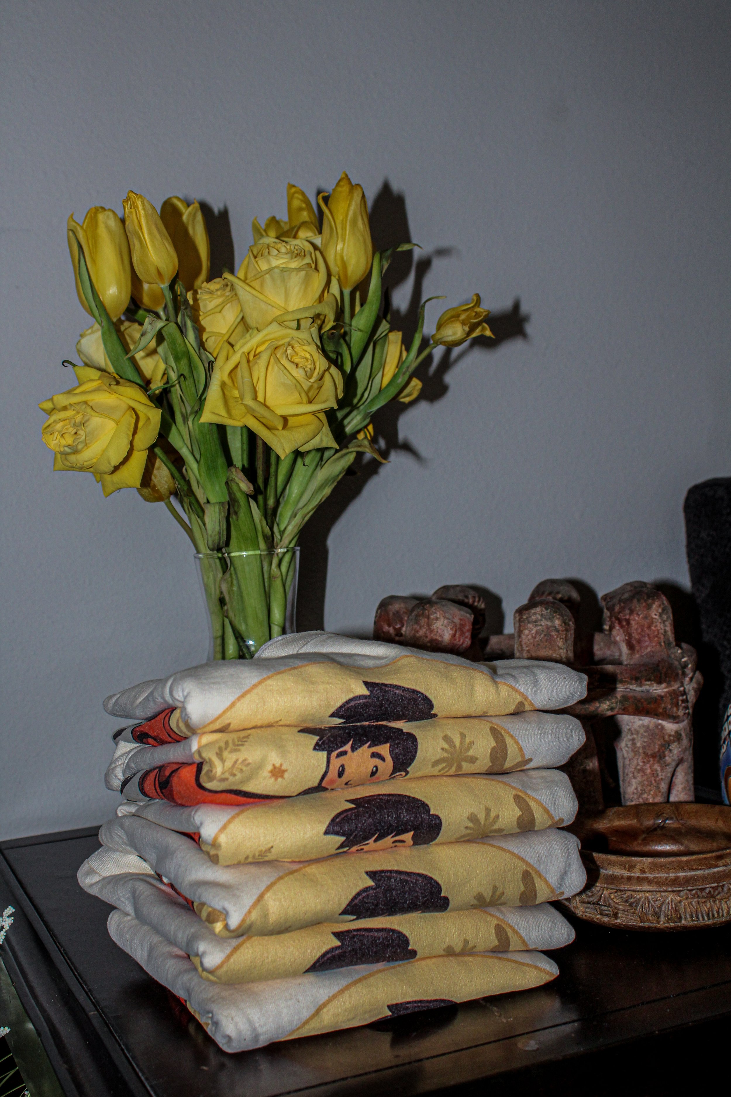 A vase of yellow roses and tulips next to a stack of folded T-shirts featuring a cartoon design on a wooden surface.