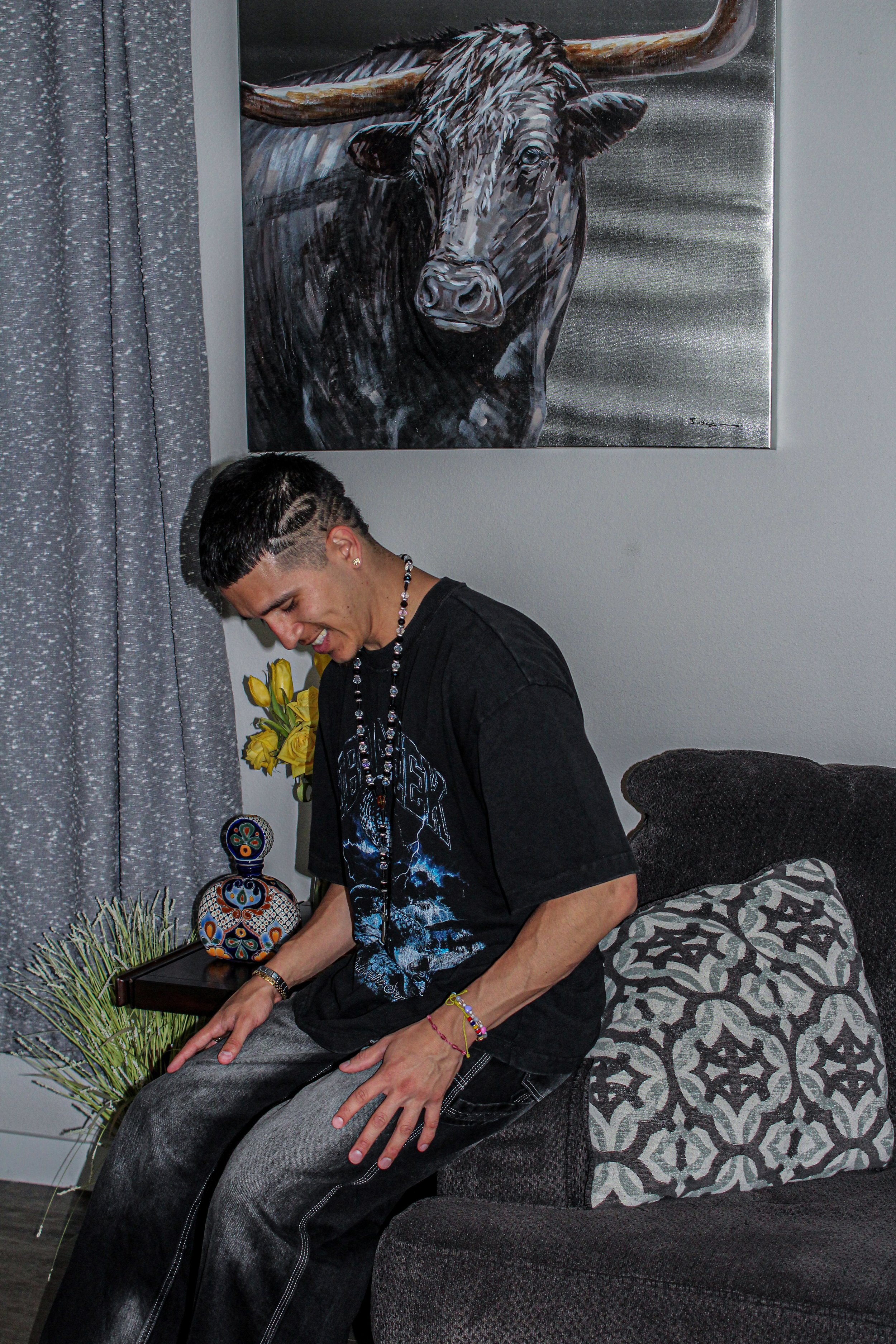 Man sitting on a couch with a painting of a bull above him, wearing a black t-shirt and jewelry, with decorative items and yellow flowers beside him.