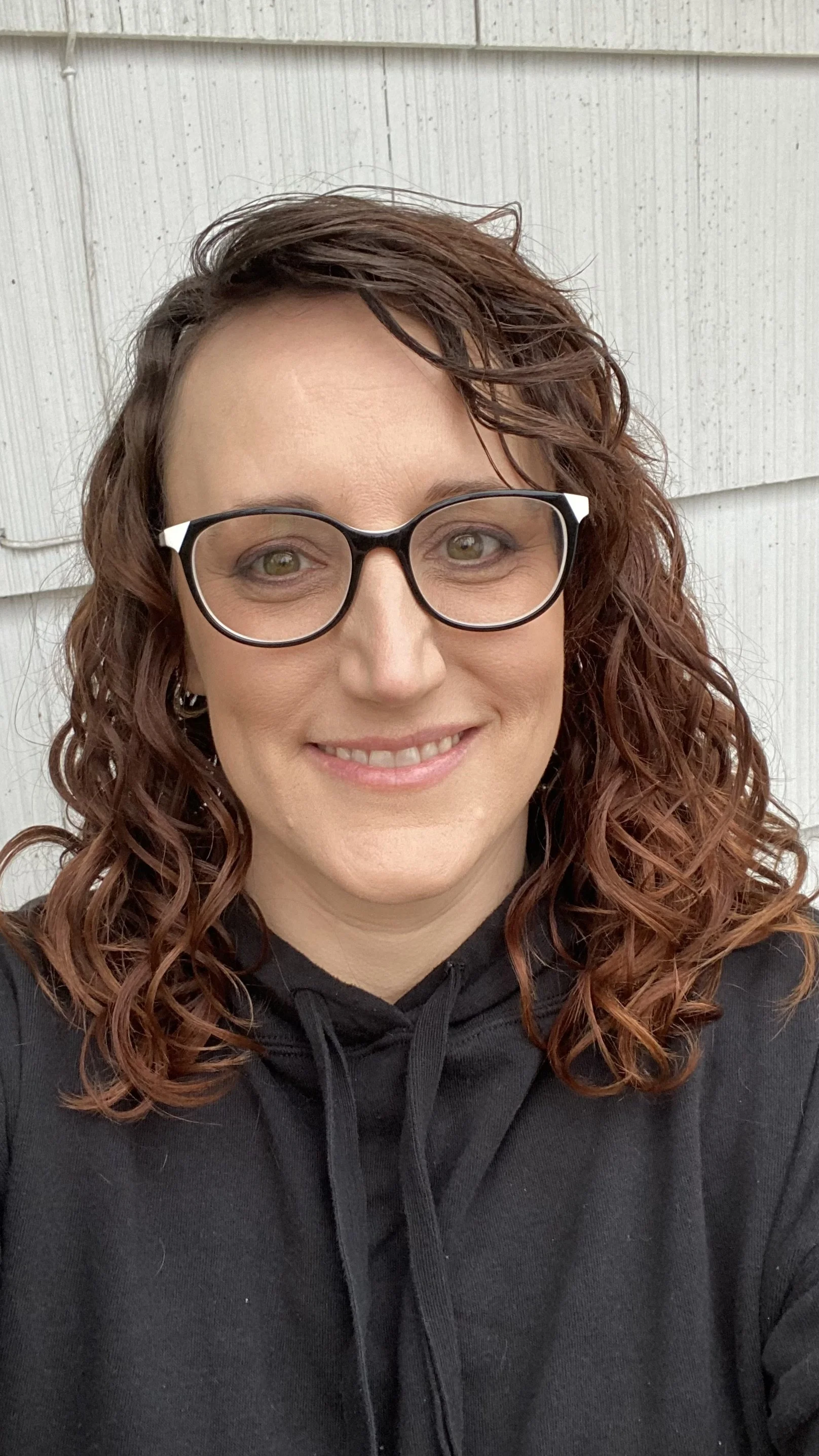 Person wearing glasses and a black hoodie, smiling in front of a light-colored wall.
