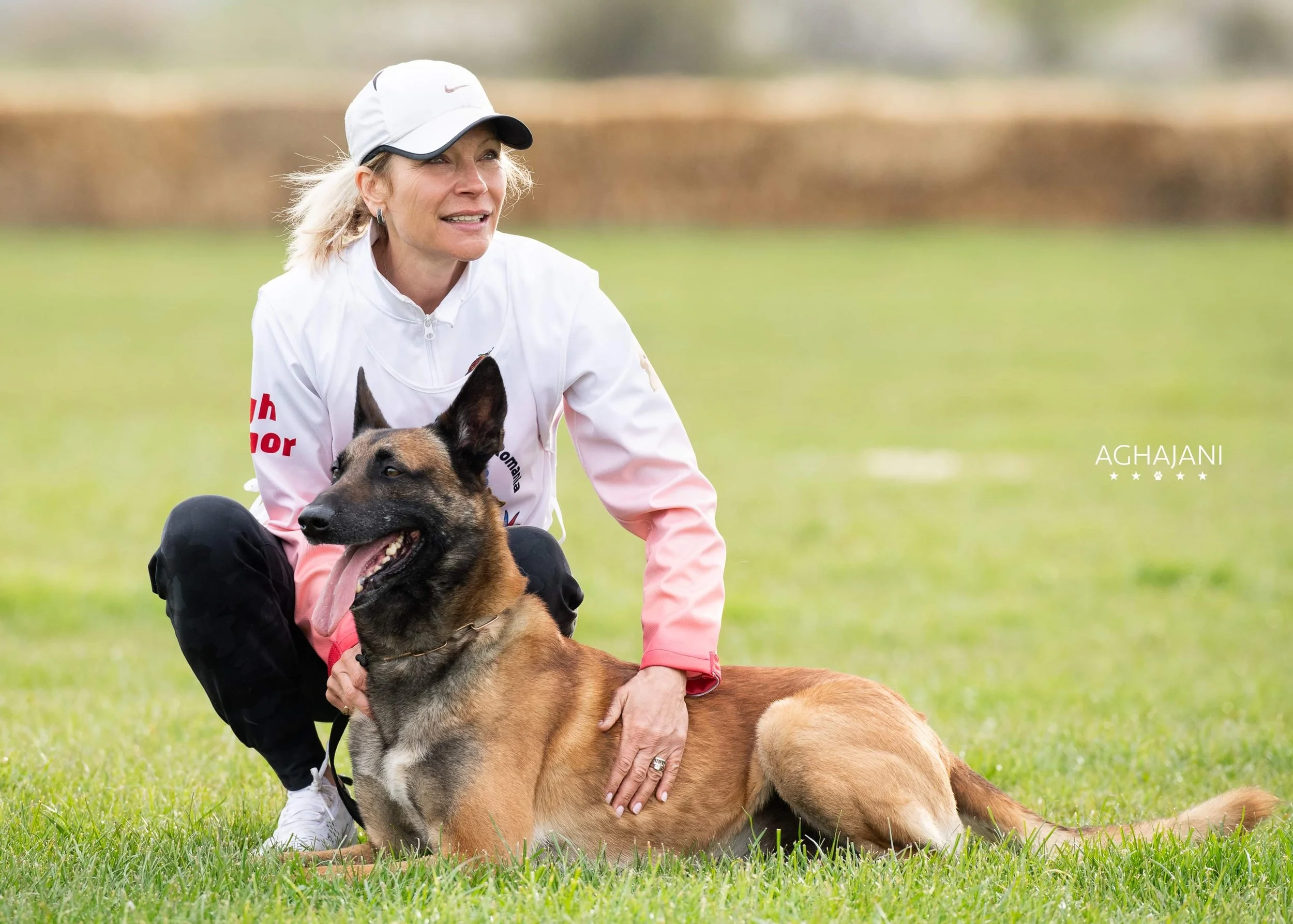 A woman with blonde hair wearing a white and pink jacket, black pants, and a white cap, kneeling on grass, holding a Belgian Malinois with a tan coat and black face, who is lying down with tongue out, in an outdoor field.