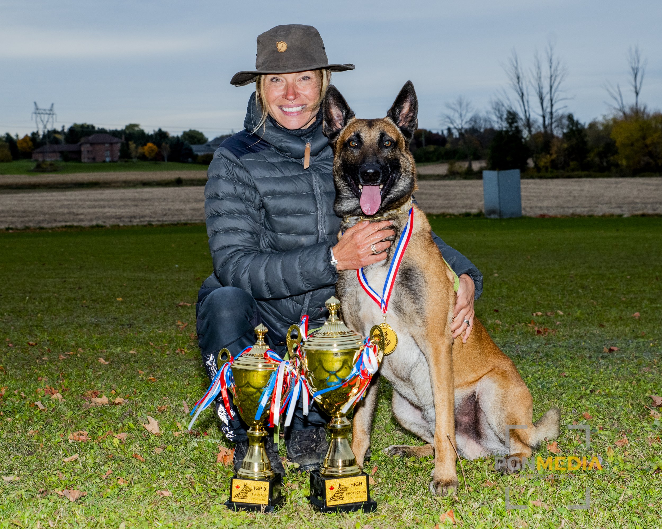 A woman in outdoor clothing, including a wide-brimmed hat, kneels on grass with a Belgian Malinois dog. Both are adorned with medals and ribbons. Three trophies are in front of them on the grass, indicating a dog competition or awards ceremony, with a rural background of fields, trees, and a cloudy sky.